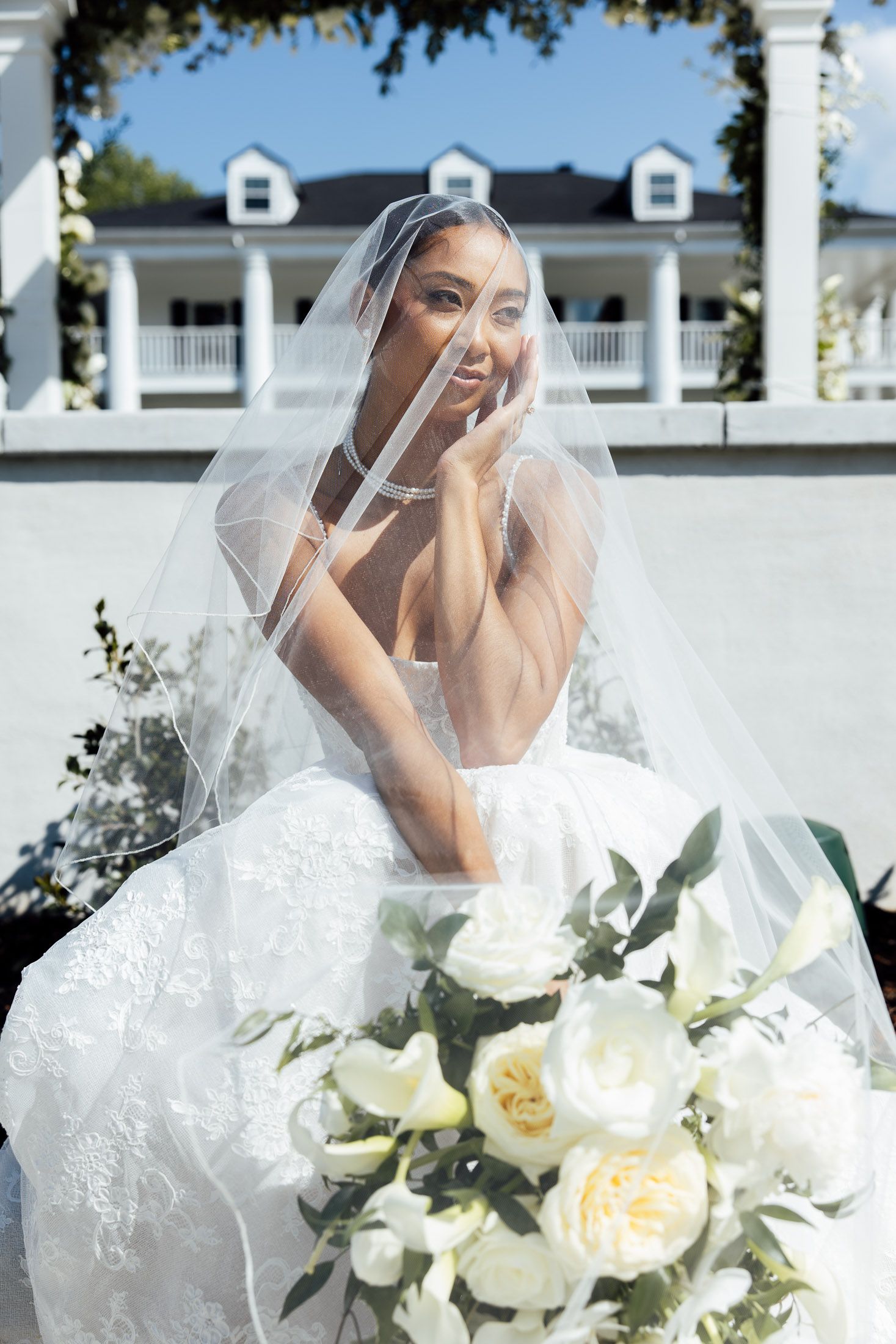 Bride in white lace gown and veil holds bouquet of white flowers outside elegant mansion on sunny day.