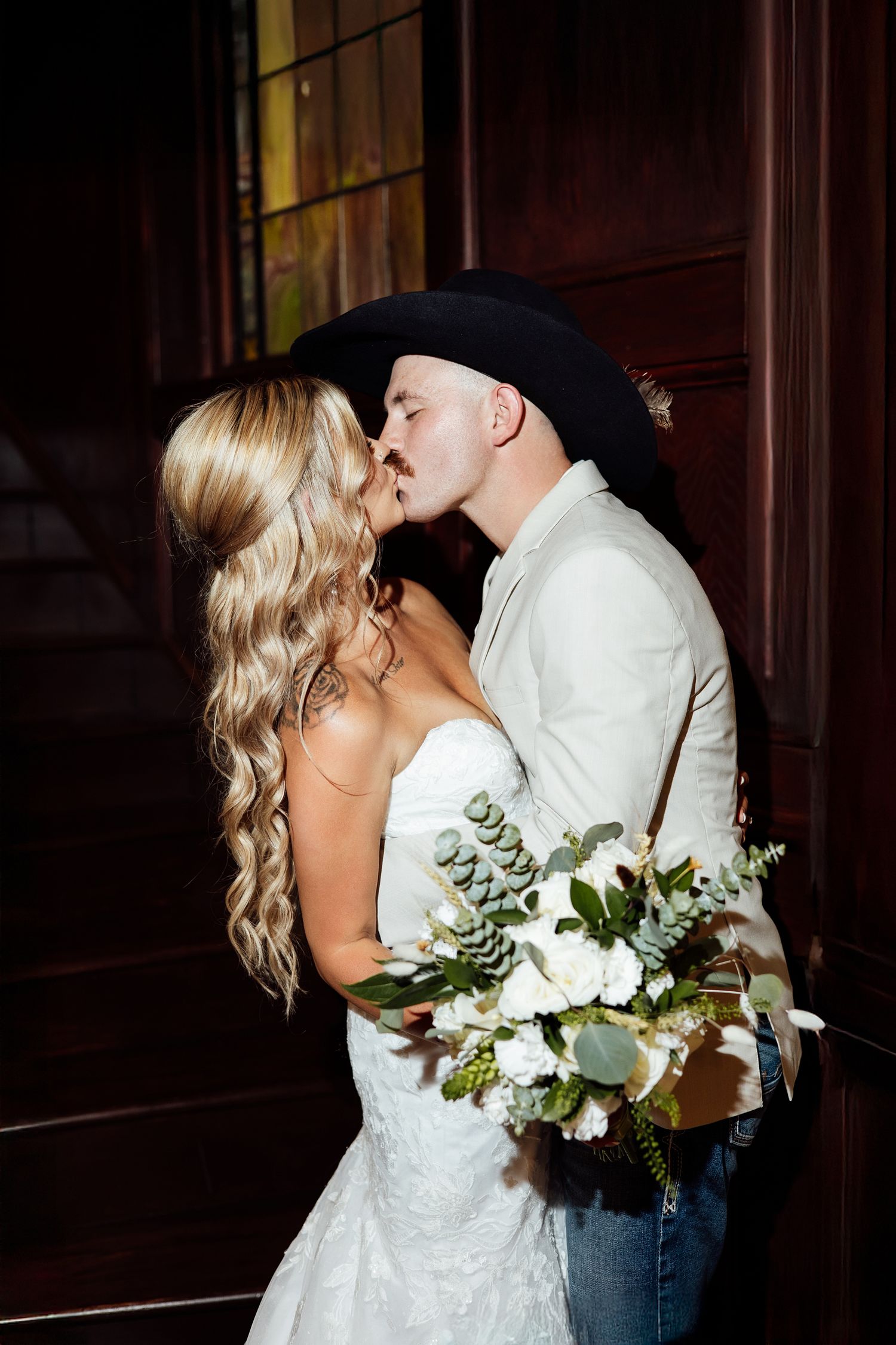 Bride and groom sharing a kiss, she holds white floral bouquet, he wears black cowboy hat and tan jacket.
