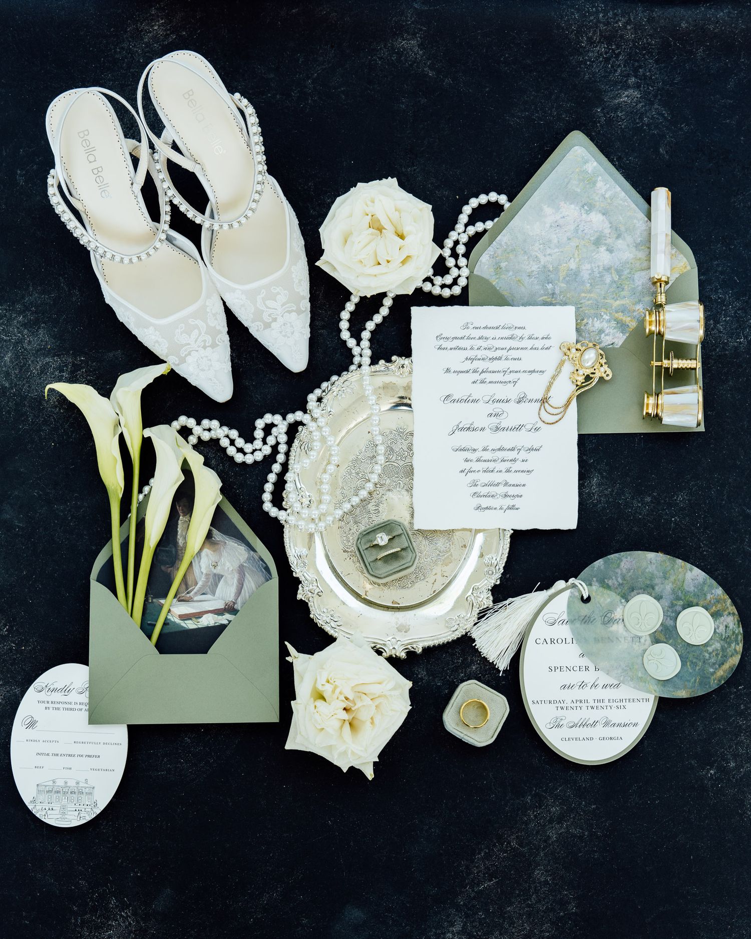 Elegant wedding flat lay with pearl heels, invitation suite, roses, calla lilies, rings, and pearl necklace on dark background.