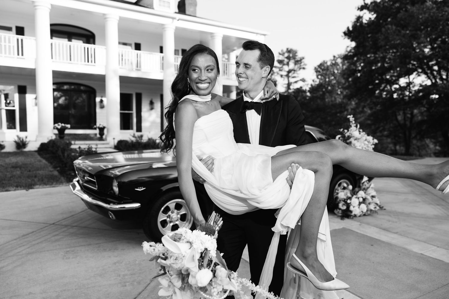 Groom carries bride in wedding attire beside a classic black car outside an elegant white mansion.