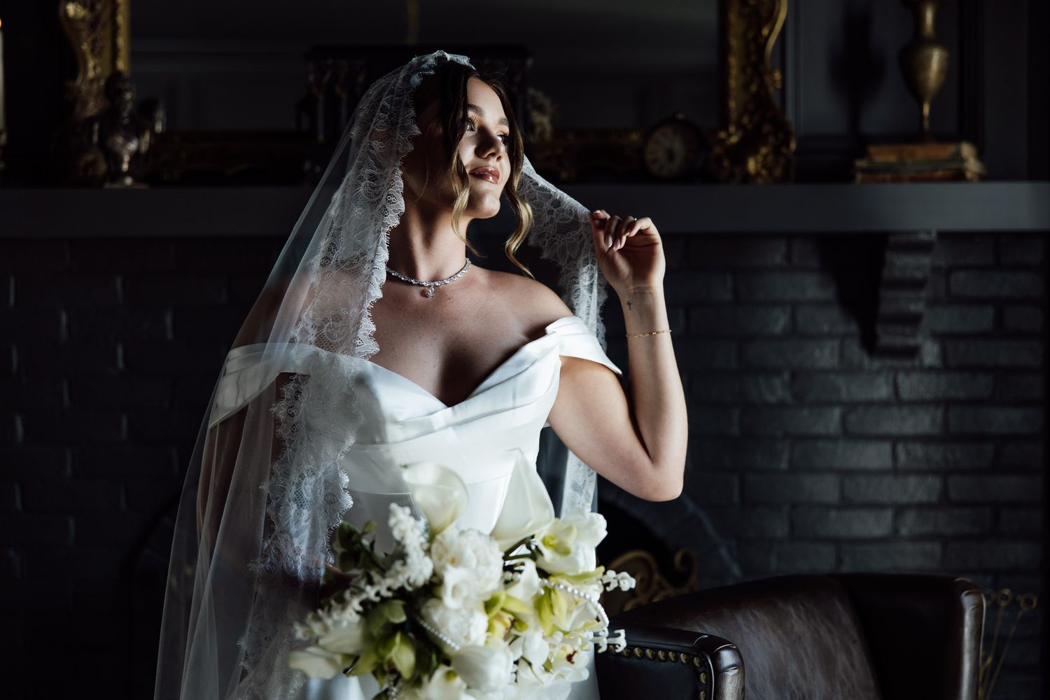 Elegant bride in white off-shoulder gown and veil seated in dark room holding bouquet of white flowers.
