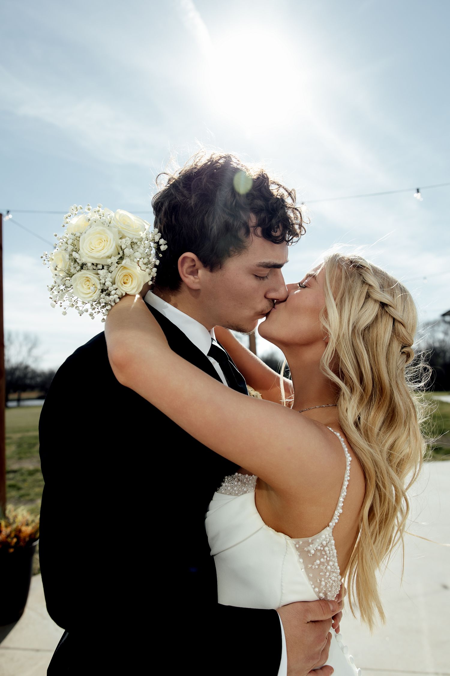 Bride and groom sharing a romantic kiss outdoors on their wedding day, with bride holding white rose bouquet in sunny weather.