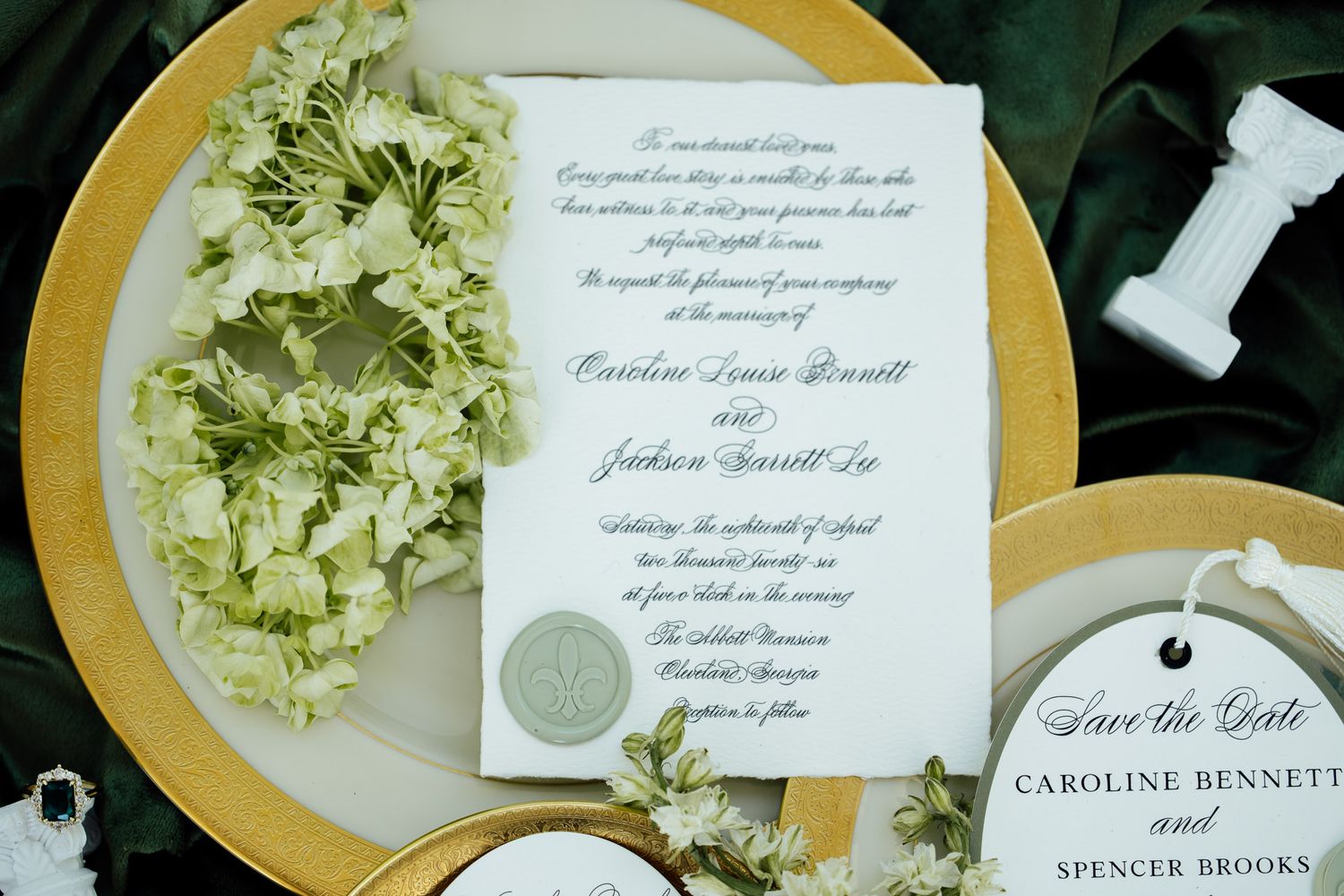 Elegant wedding invitation suite with wax seal on gold charger plate, surrounded by white hydrangea floral wreath.