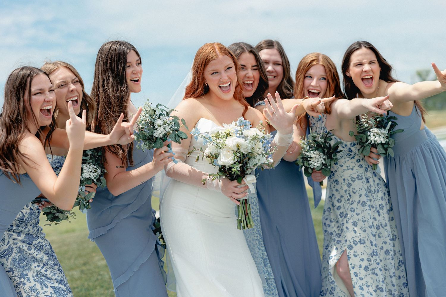 Bride in white gown holds bouquet as bridesmaids in blue dresses excitedly point and celebrate outdoors.