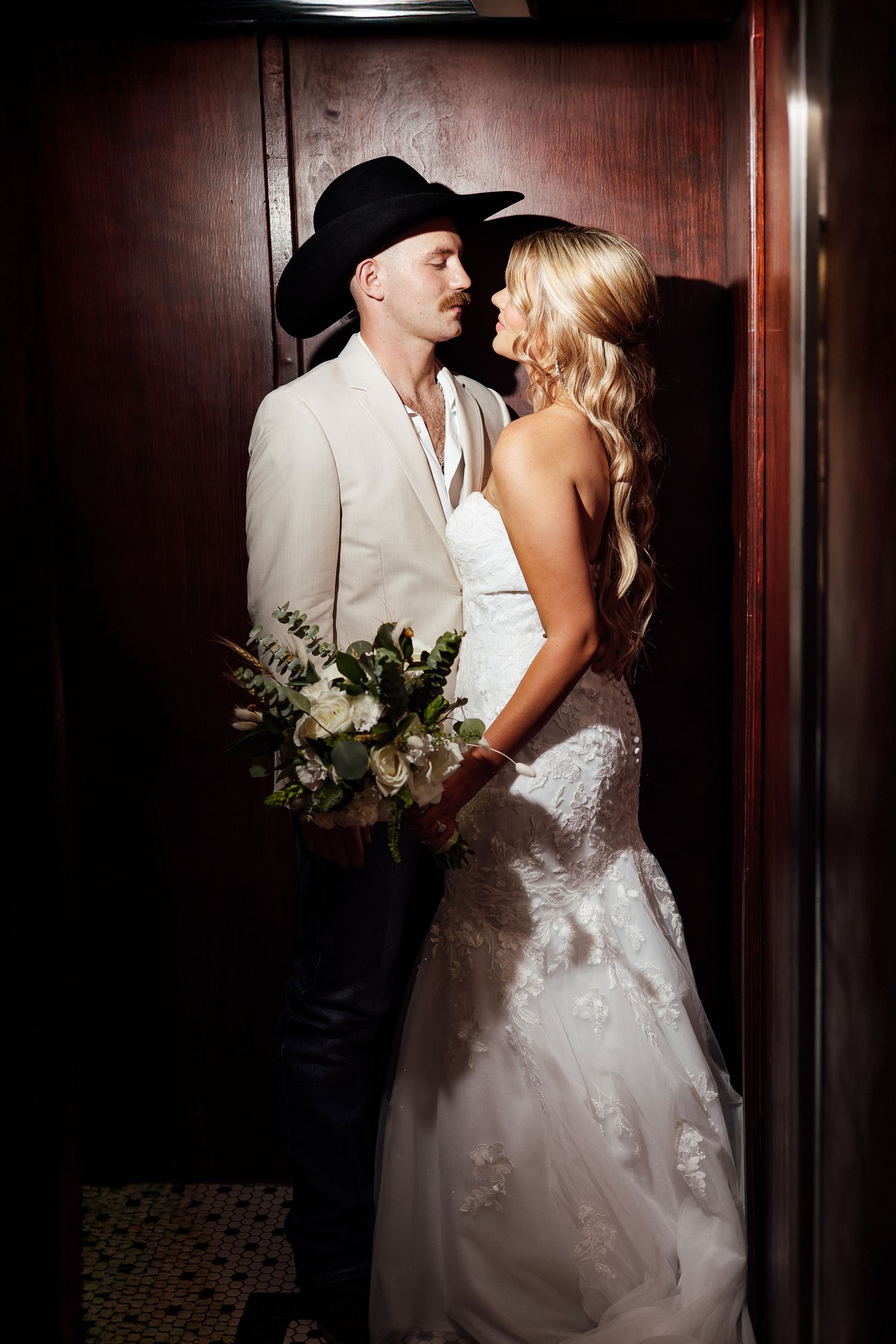 A bride and groom share a romantic moment in an elevator, she in lace gown, he in cream suit and black cowboy hat.