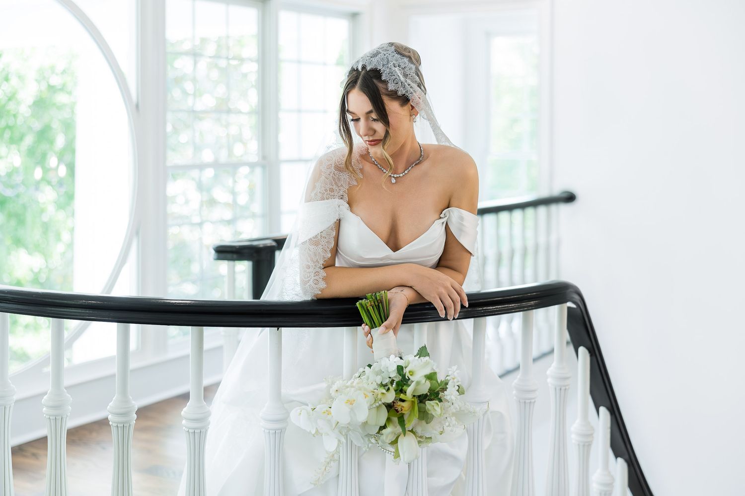 Bride in white off-shoulder gown with veil holds bouquet while leaning on staircase railing in bright elegant venue.