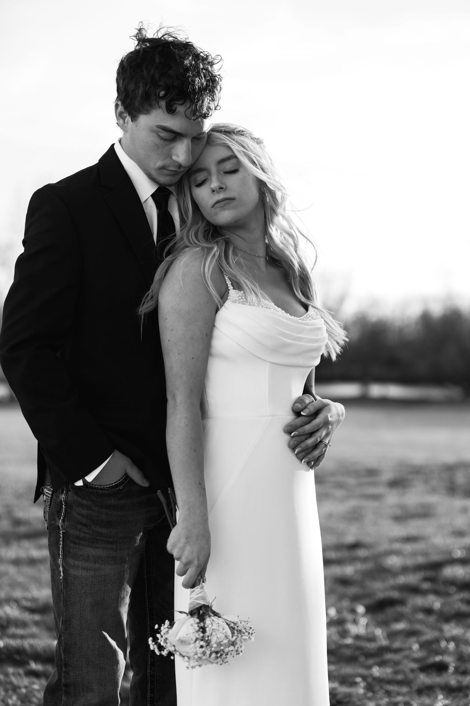 Black and white photo of a bride and groom embracing outdoors, she holds a small bouquet in a romantic pose.