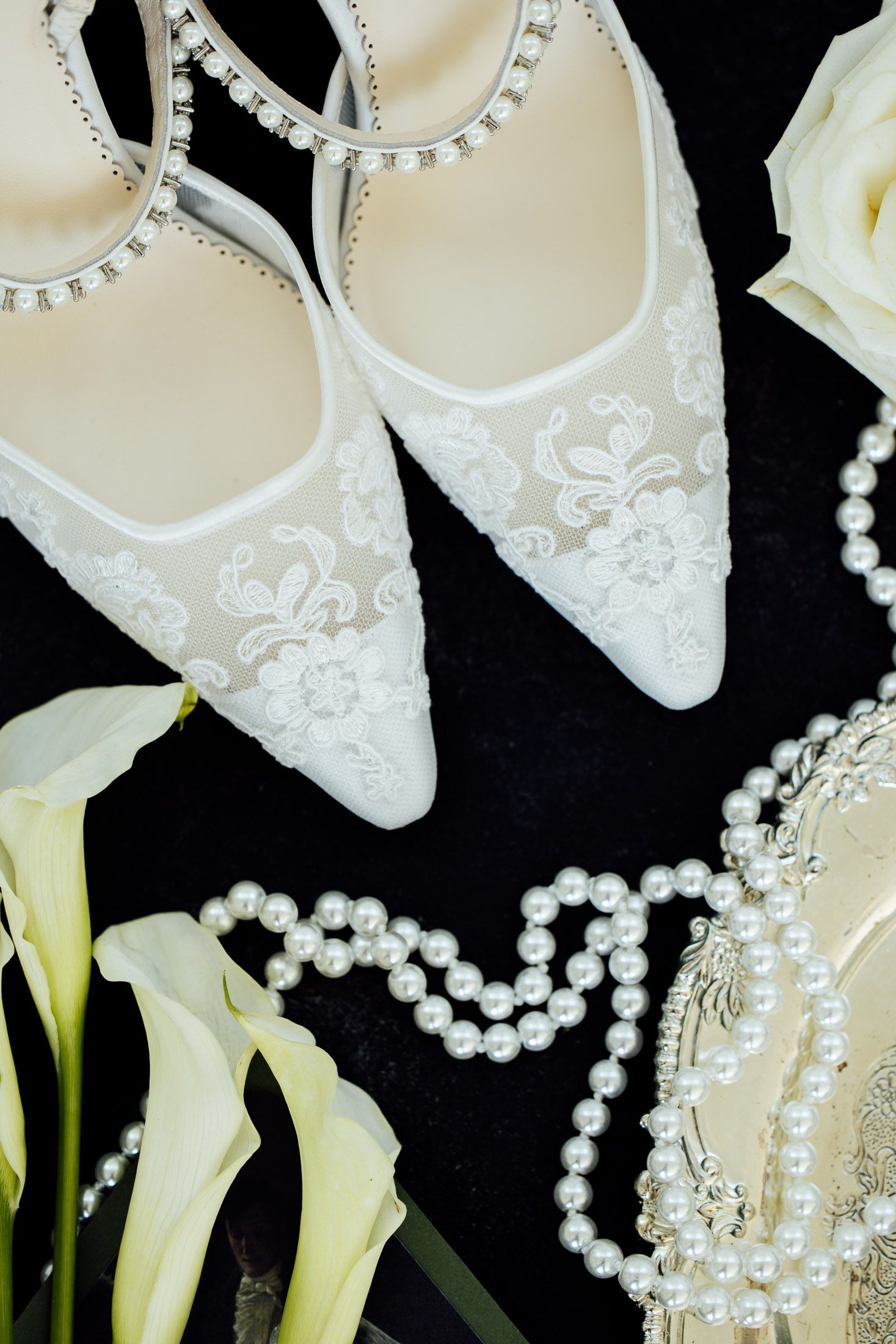 White lace bridal heels with crystal ankle straps, pearl necklace, and calla lilies on dark background.