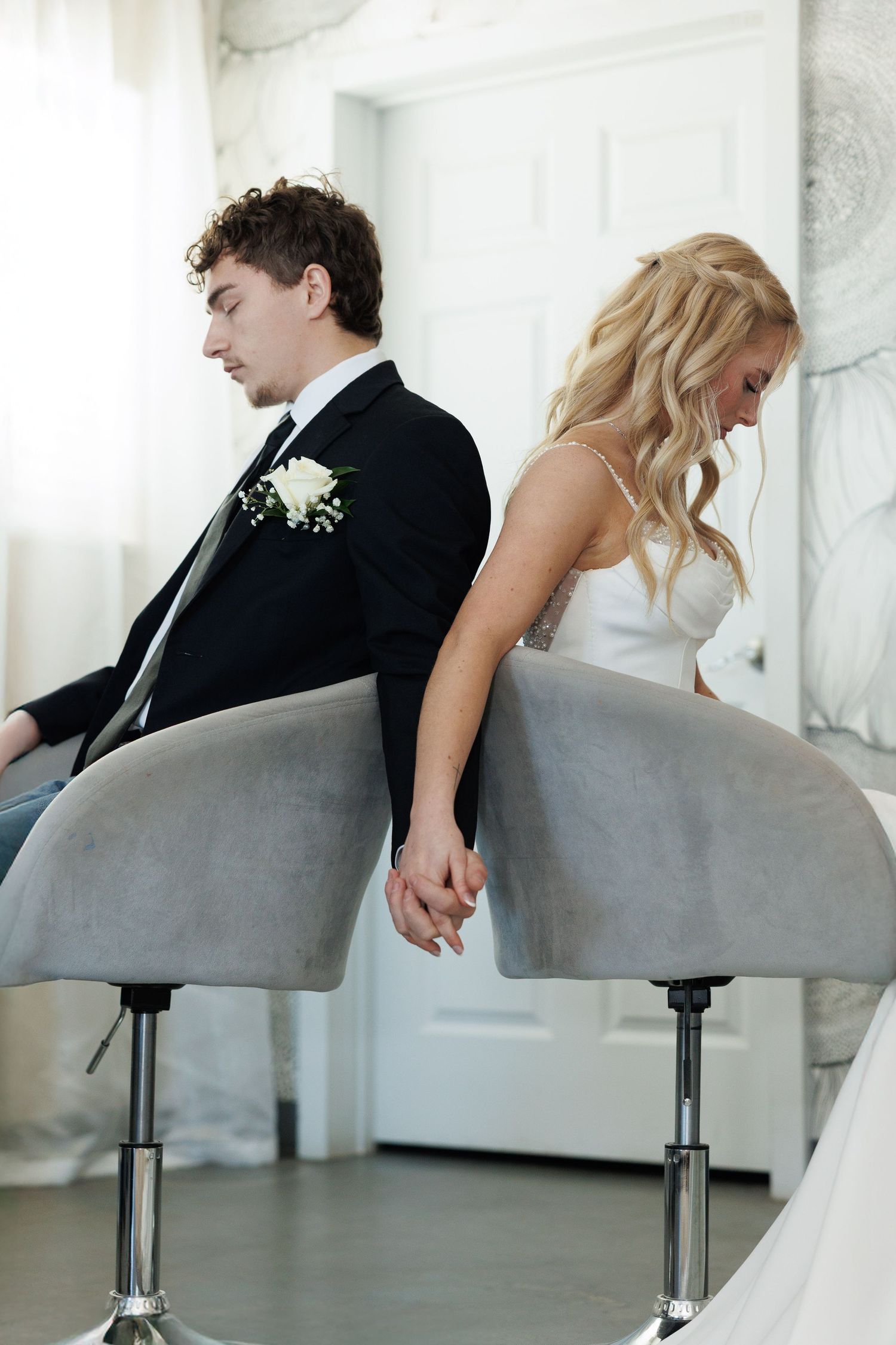 Bride and groom sit back-to-back on gray chairs, holding hands tenderly during a first look at a wedding