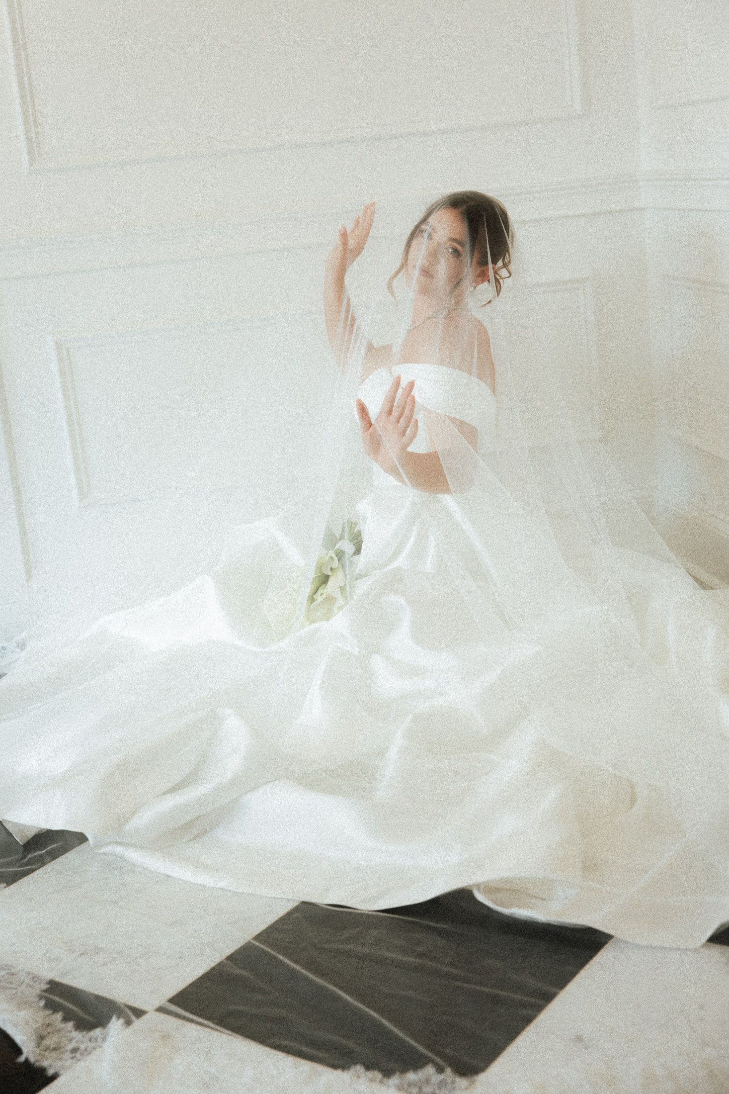 A bride in an elegant off-shoulder white ball gown sits gracefully, her veil flowing around her in a bright, airy room.