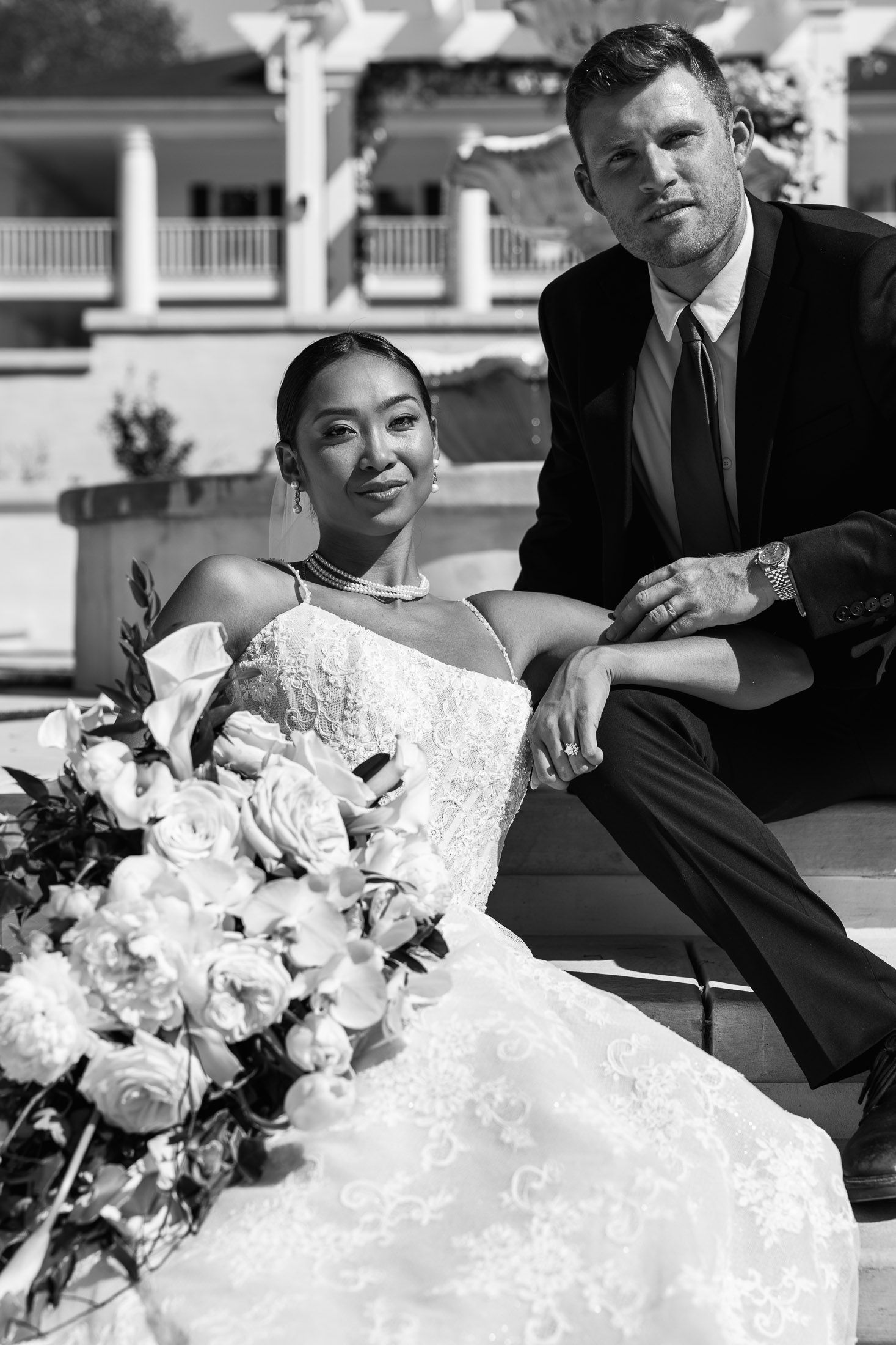 Black and white wedding portrait of bride in lace gown with bouquet and groom in suit posing outdoors.