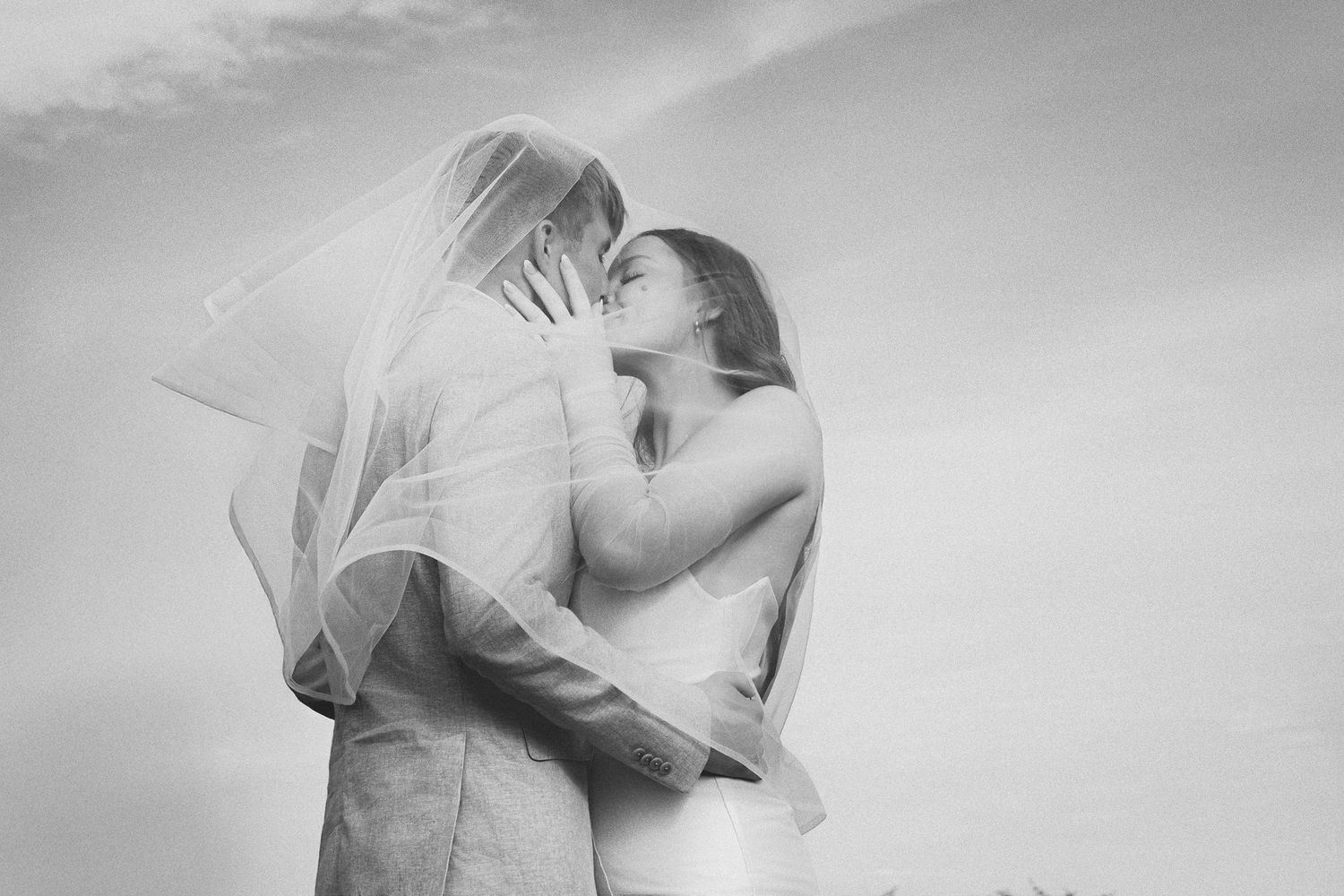 Black and white photo of a bride and groom sharing a romantic kiss under a flowing veil against a soft sky.