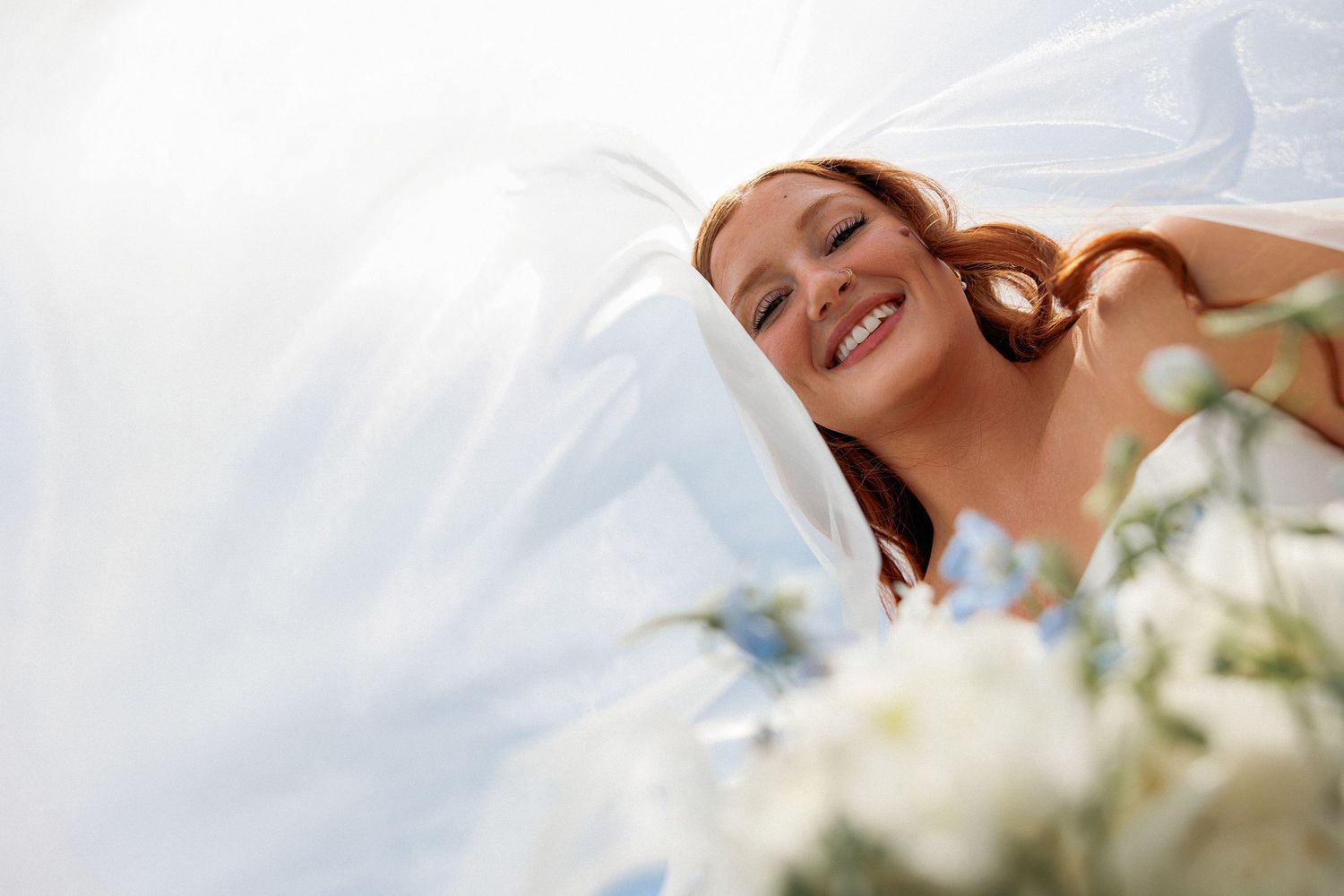 Smiling redhead bride in white veil holding blue and white bouquet, photographed from below against bright sky.