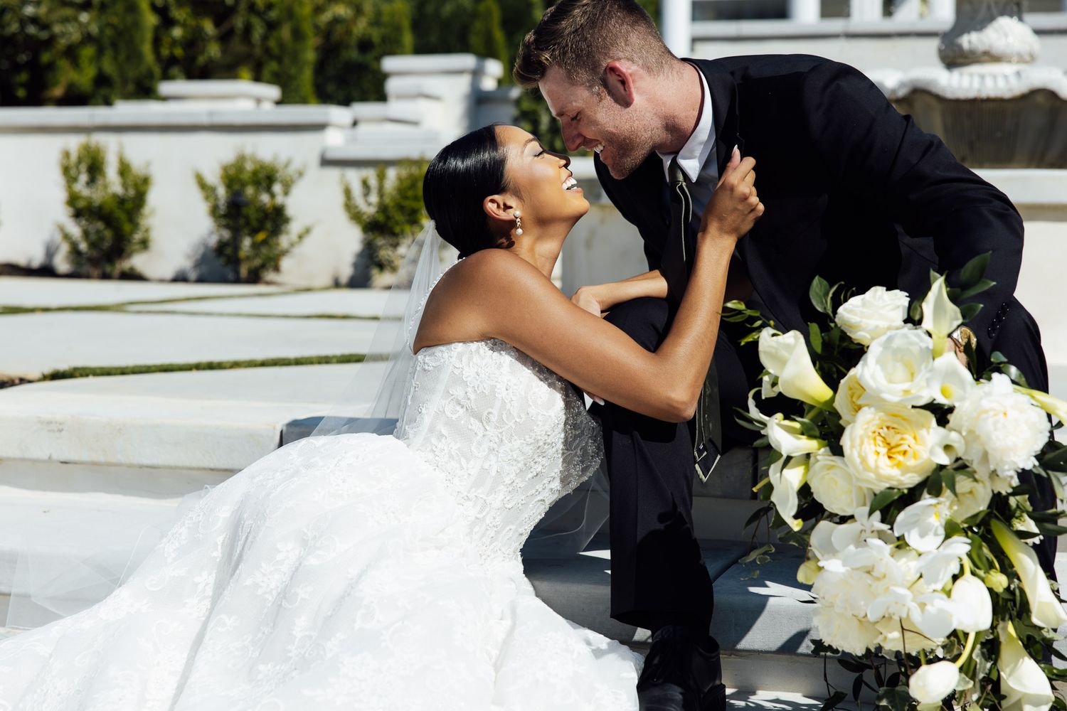 A bride and groom share a romantic moment, with white floral bouquet, on outdoor steps during their wedding day.