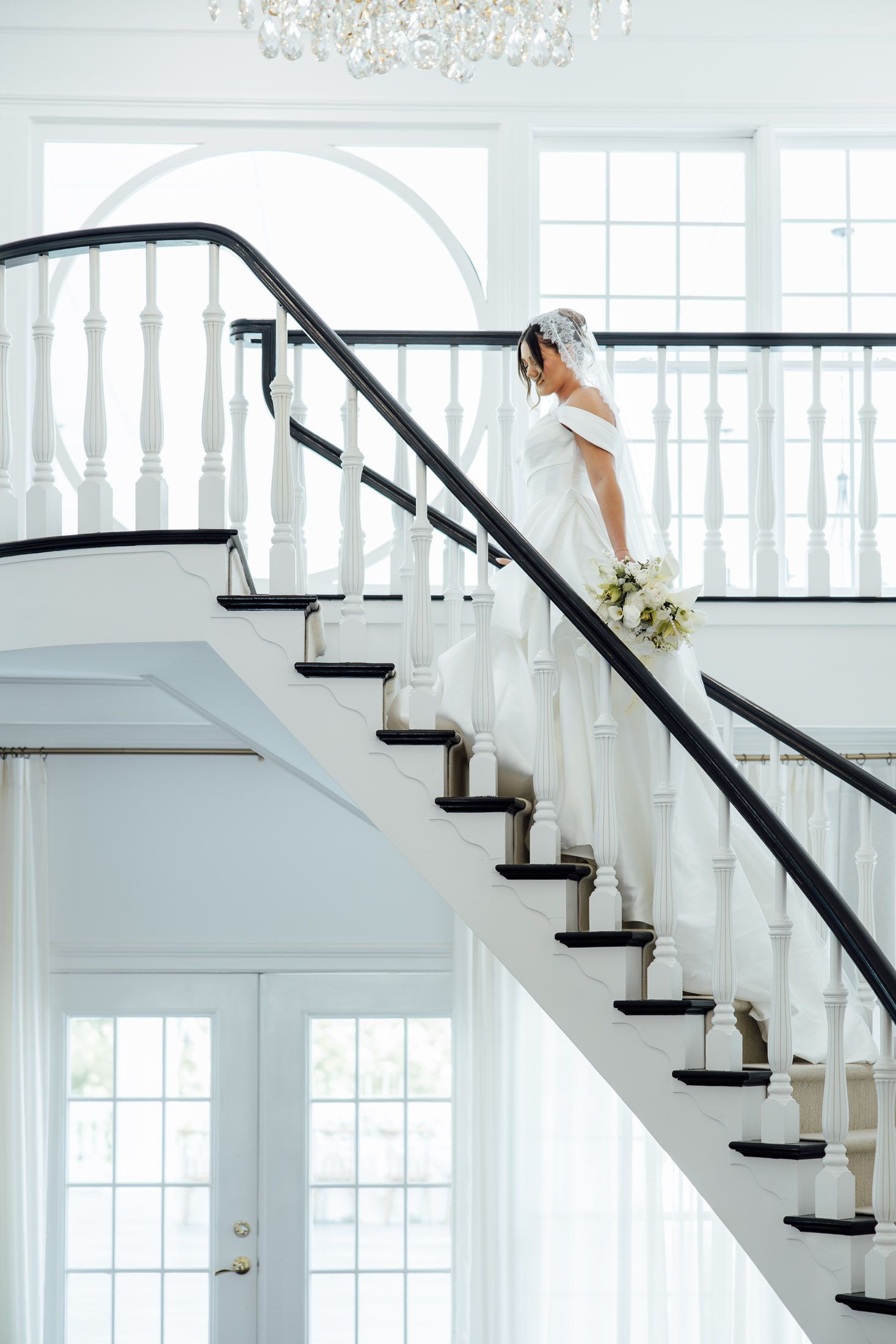 Bride in white gown holding bouquet descends elegant staircase with black railings in bright white venue.