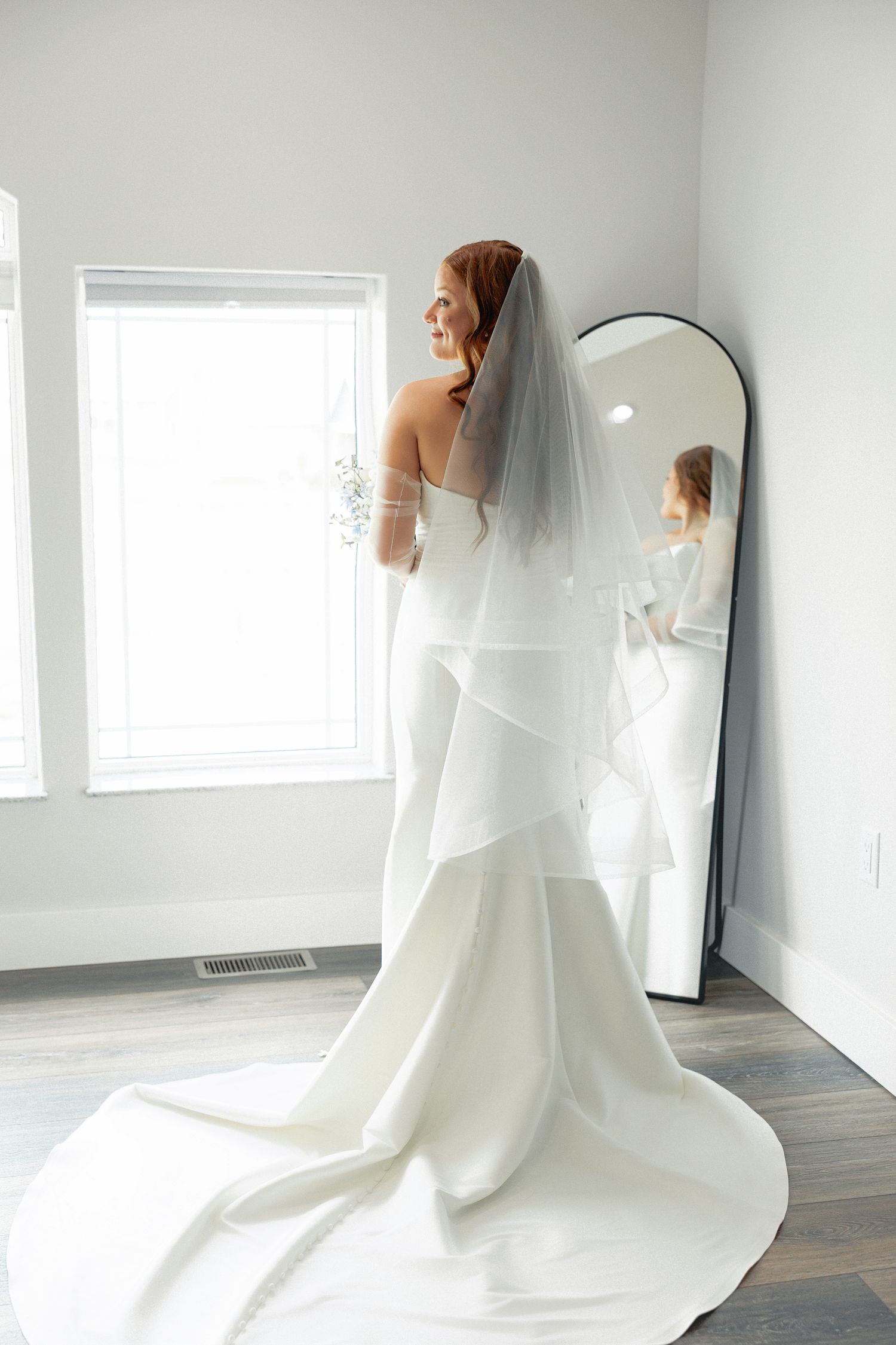 Bride in white mermaid gown with veil stands by window near floor mirror in bright bridal suite.