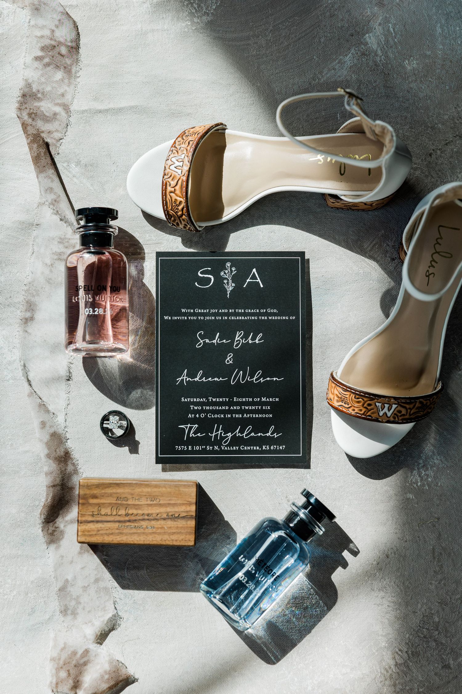 Wedding flat lay with dark invitation, white heeled sandals, perfume bottles, and wooden block on marble surface.