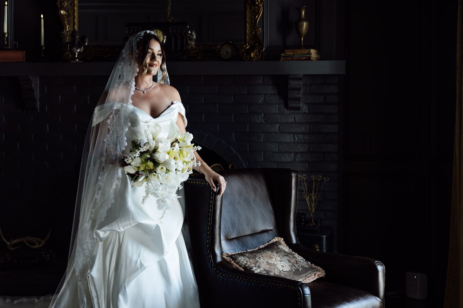 Bride in white off-shoulder gown and veil holds white bouquet in dark elegant room with fireplace.