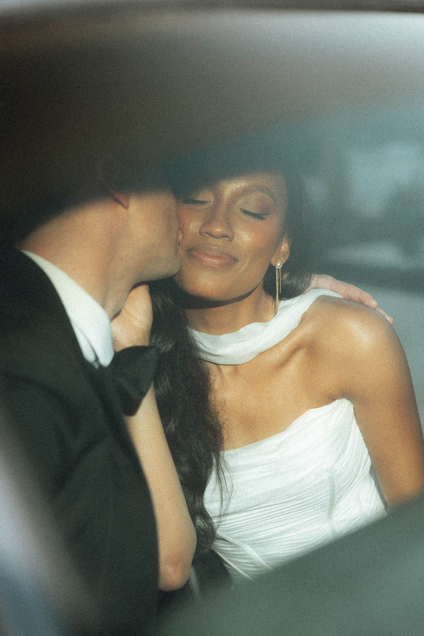 A groom in a black tuxedo kisses his bride on the cheek as she smiles in a white dress inside a car.