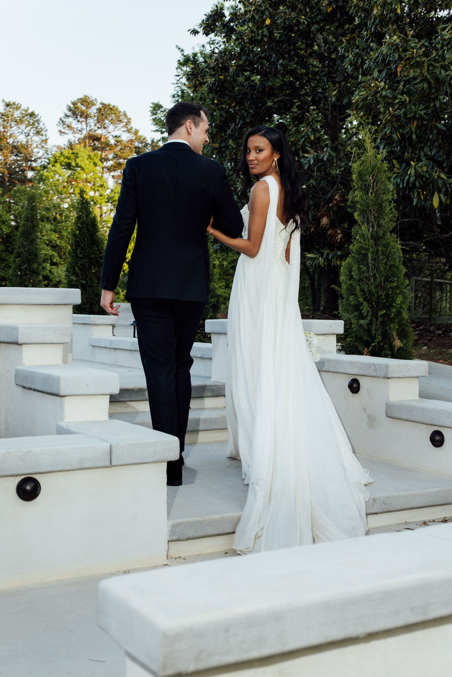 A bride in a flowing white gown and groom in black suit walk up white stone steps in a lush garden setting.