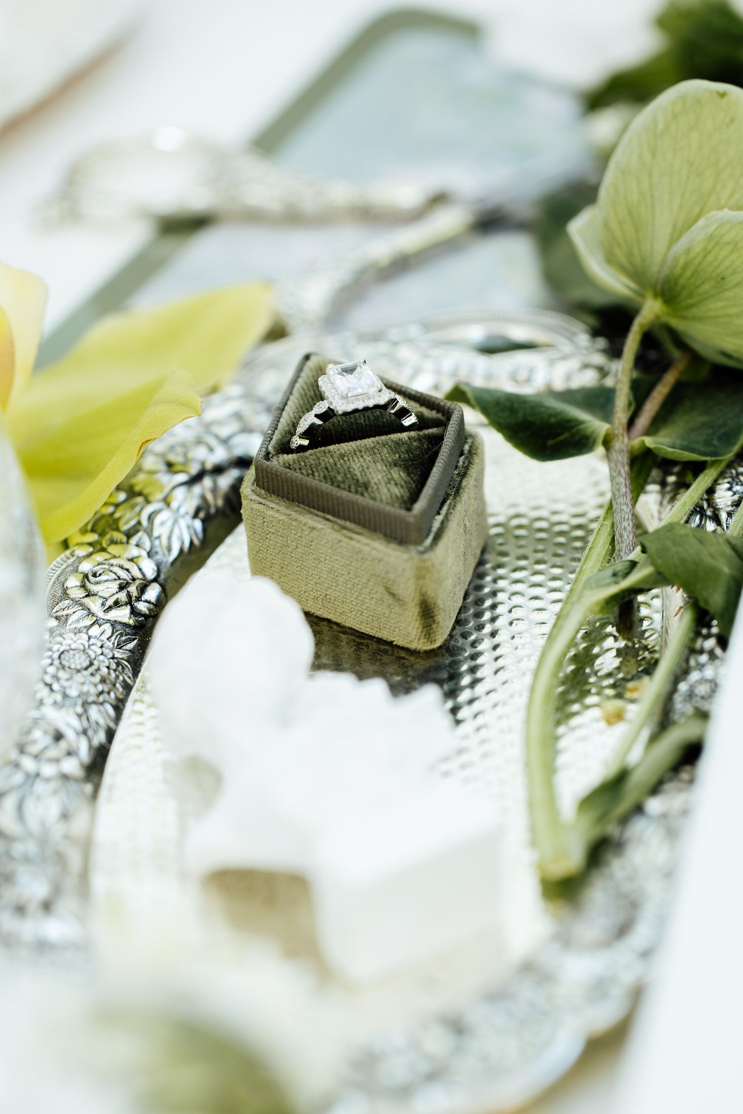 Diamond engagement ring in green velvet box on ornate silver tray with white flowers and green leaves.