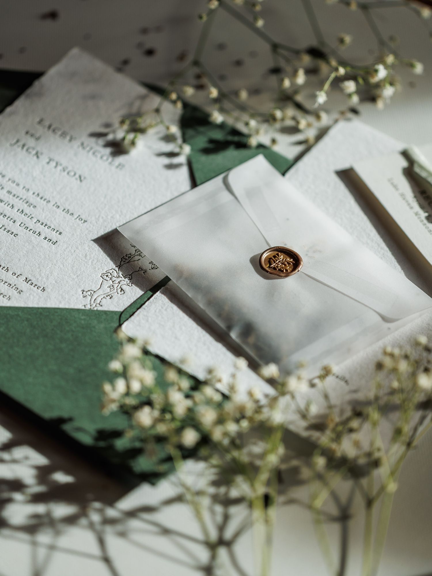 Elegant wedding invitation suite with green envelopes, vellum wrap, gold wax seal, and delicate white flowers on a dark background.
