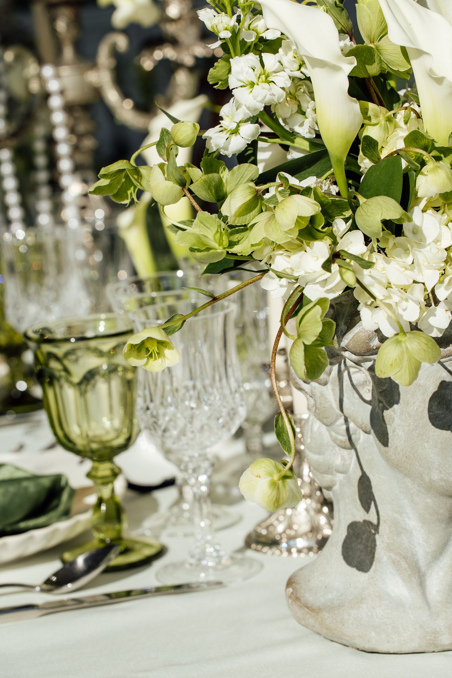 Elegant wedding table setting with white and green floral arrangement in stone vase, crystal glasses, and silver accents.