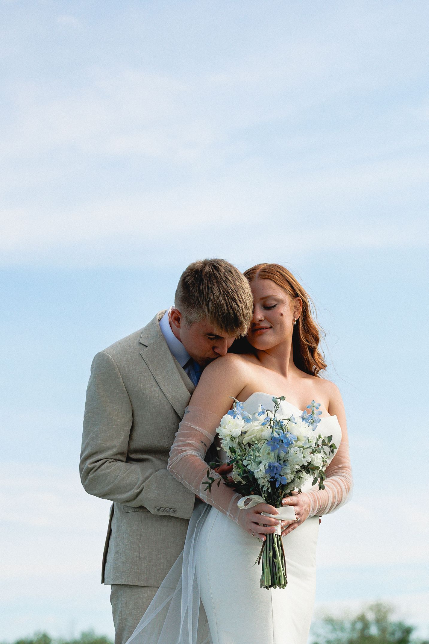 Bride and groom share a tender moment outdoors, bride holding blue and white bouquet in white gown, groom in gray suit.