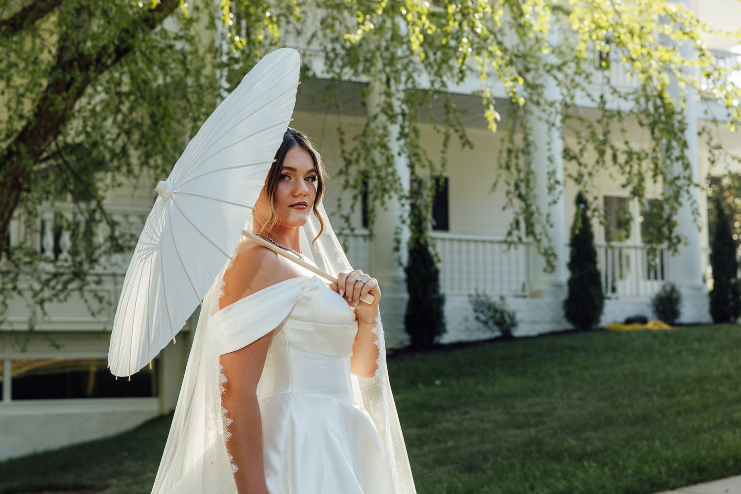 Bride in off-shoulder white gown holds parasol umbrella outdoors near white house with greenery.
