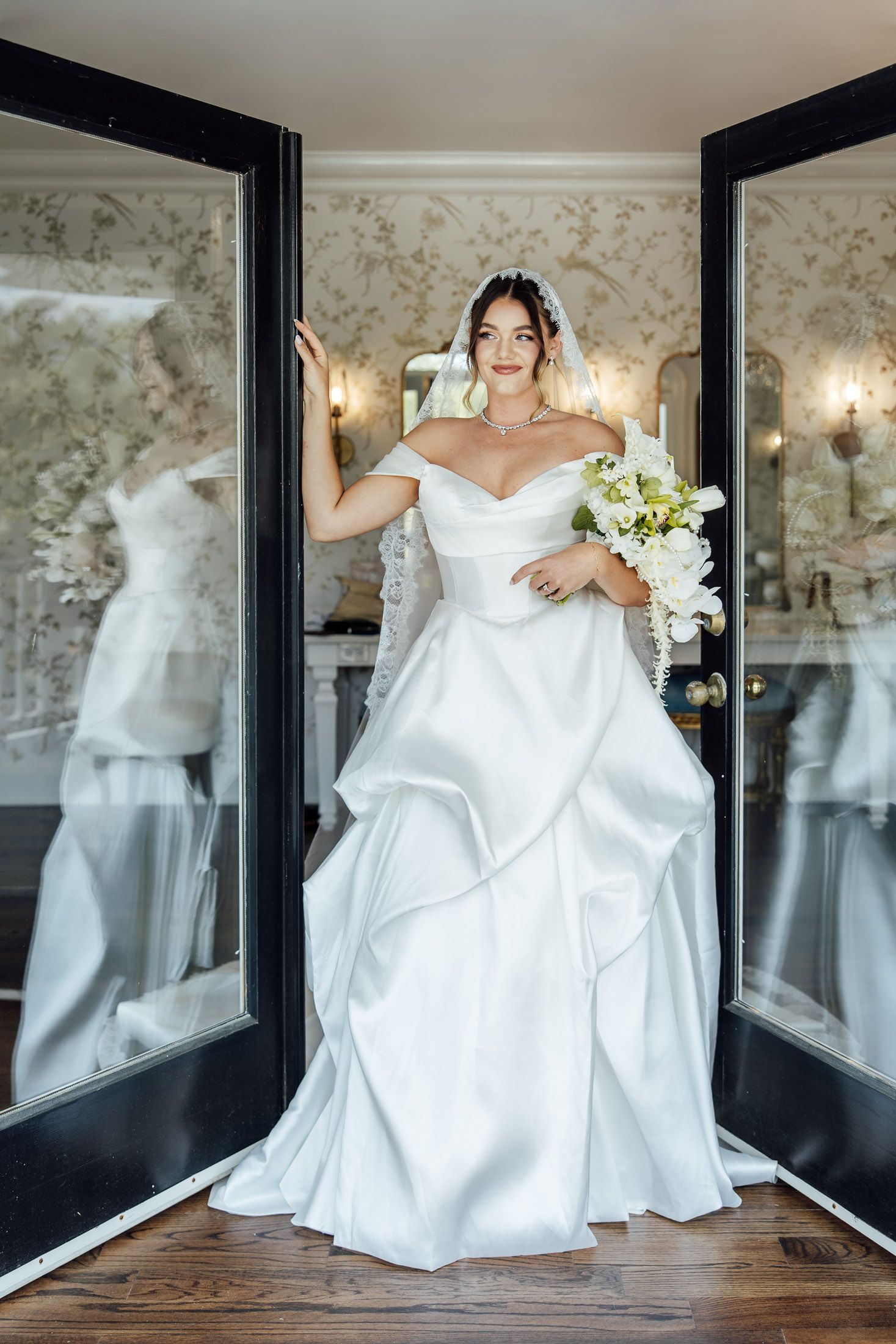 Bride in off-shoulder white satin ball gown with veil holds floral bouquet standing in elegant doorway.