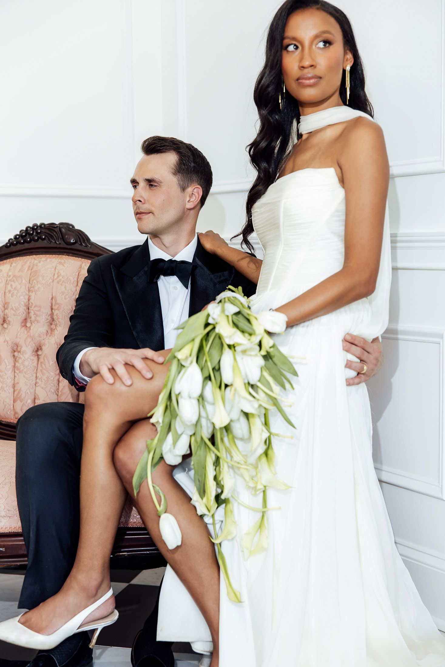 Elegant interracial wedding couple, bride in white gown holding tulip bouquet, groom in tuxedo seated on vintage chair.