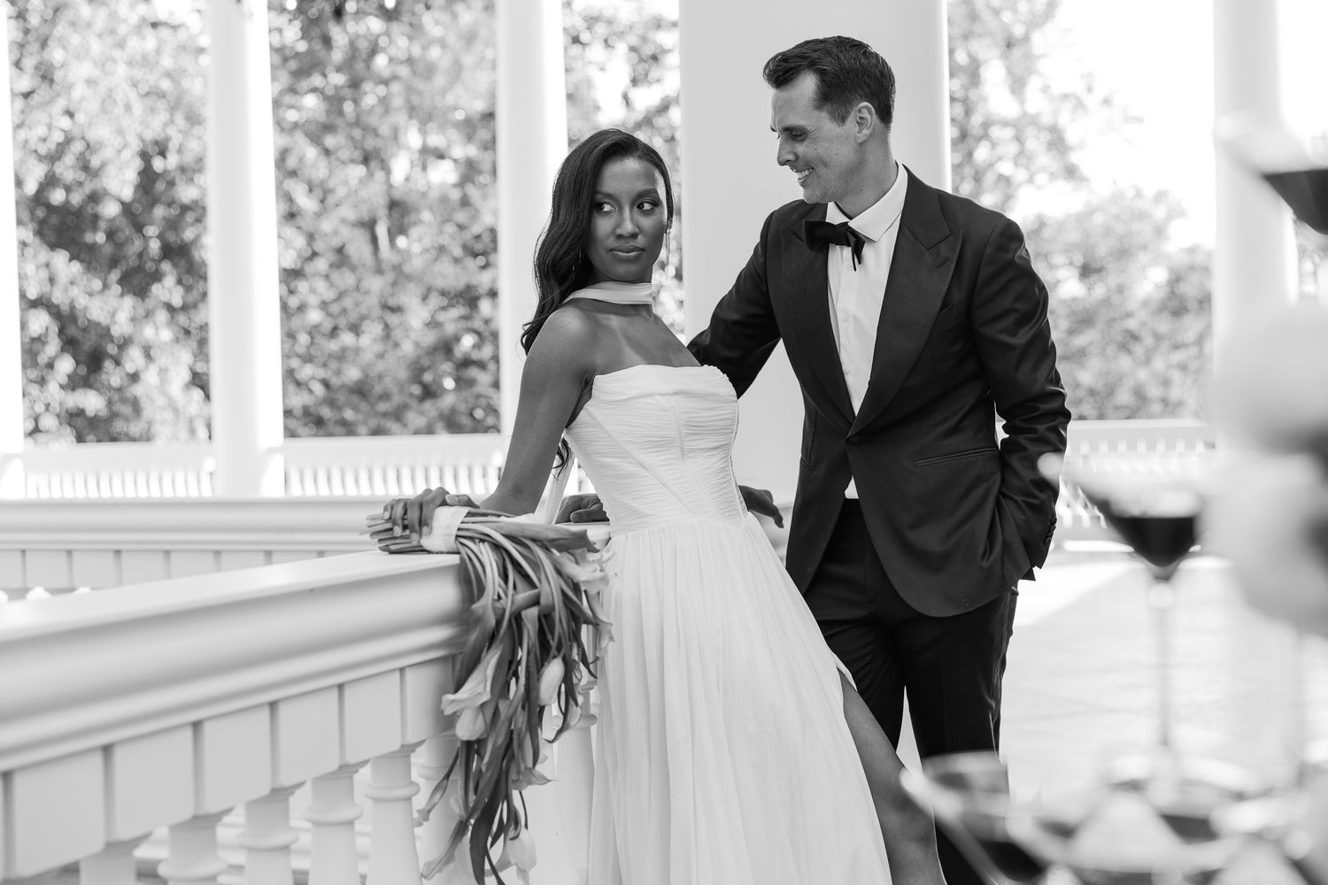 Black and white photo of an interracial couple in wedding attire posing elegantly on a white columned veranda.