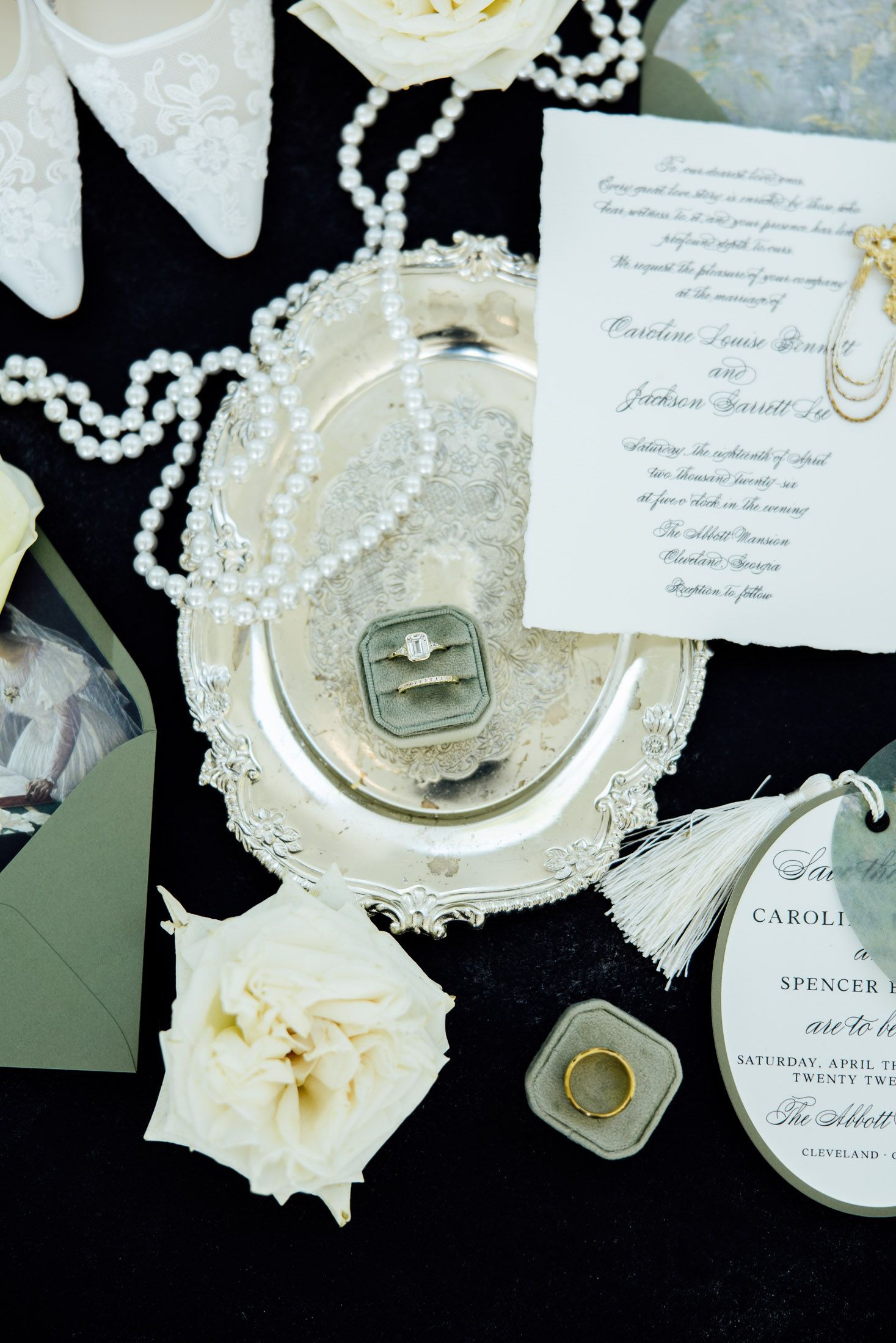 Elegant wedding flat lay with diamond ring on silver tray, pearl necklace, white roses, invitations, and gold band on black background.