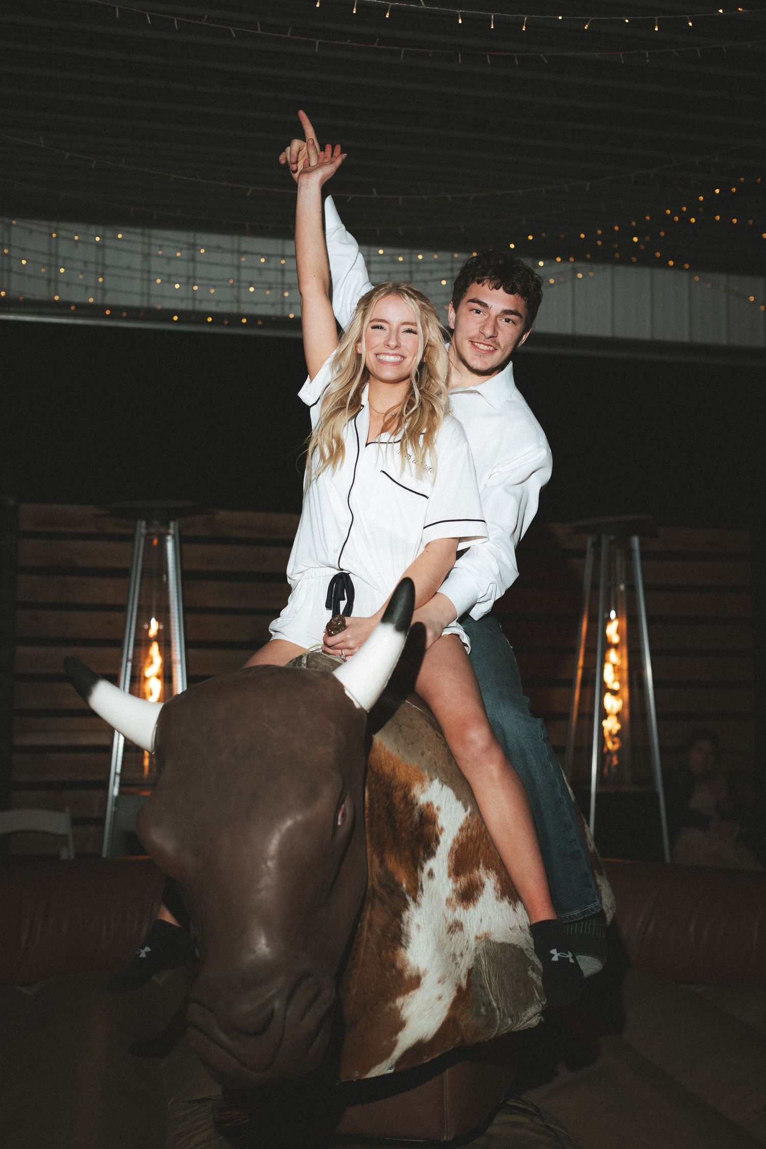 A couple rides a mechanical bull together at an indoor event, with the woman raising her arm triumphantly under warm string lights.