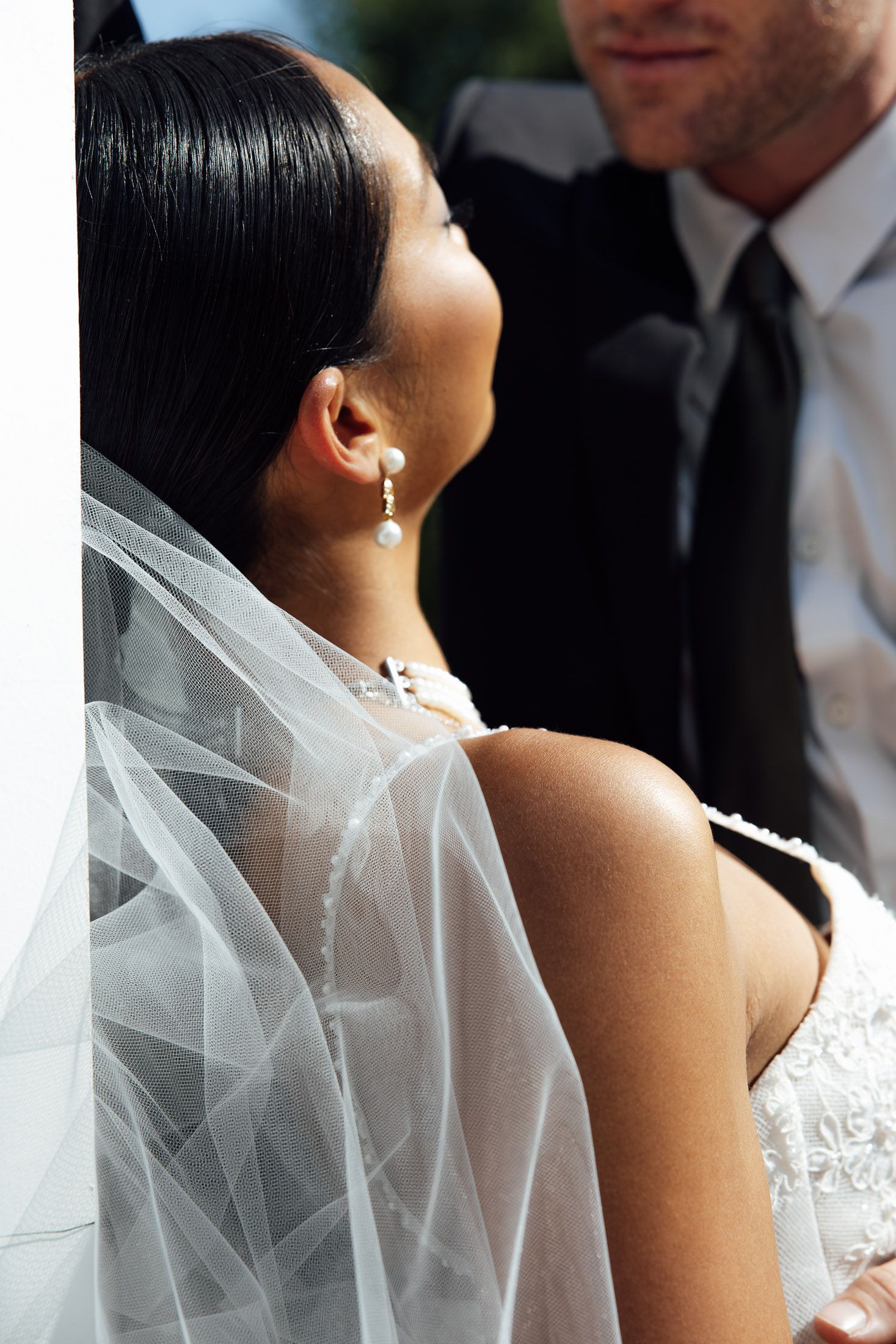 Bride in white lace gown and veil with pearl earrings looks up at groom in suit during outdoor wedding ceremony.