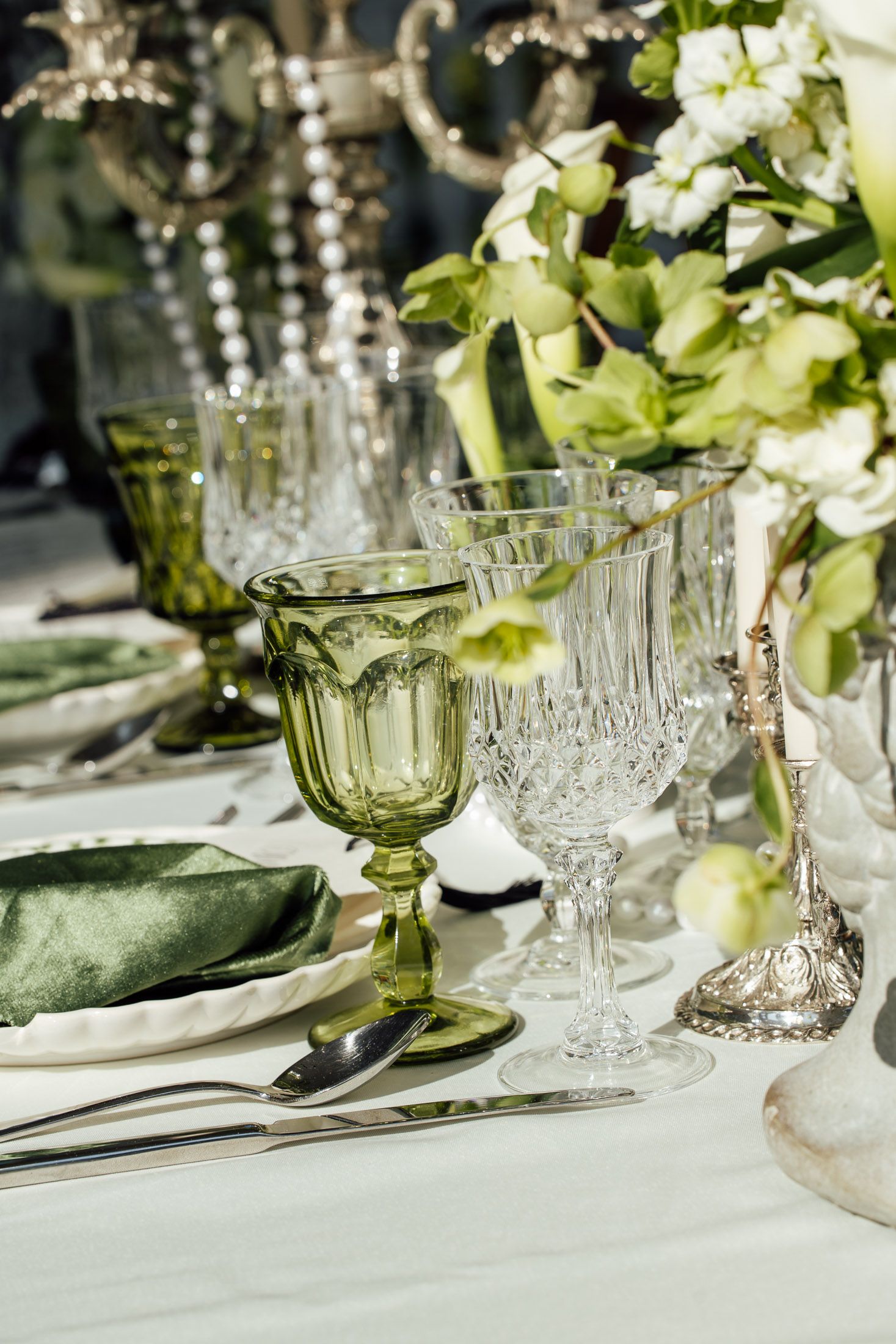 Elegant green and crystal glassware on a white wedding table setting with floral centerpiece and silver cutlery.