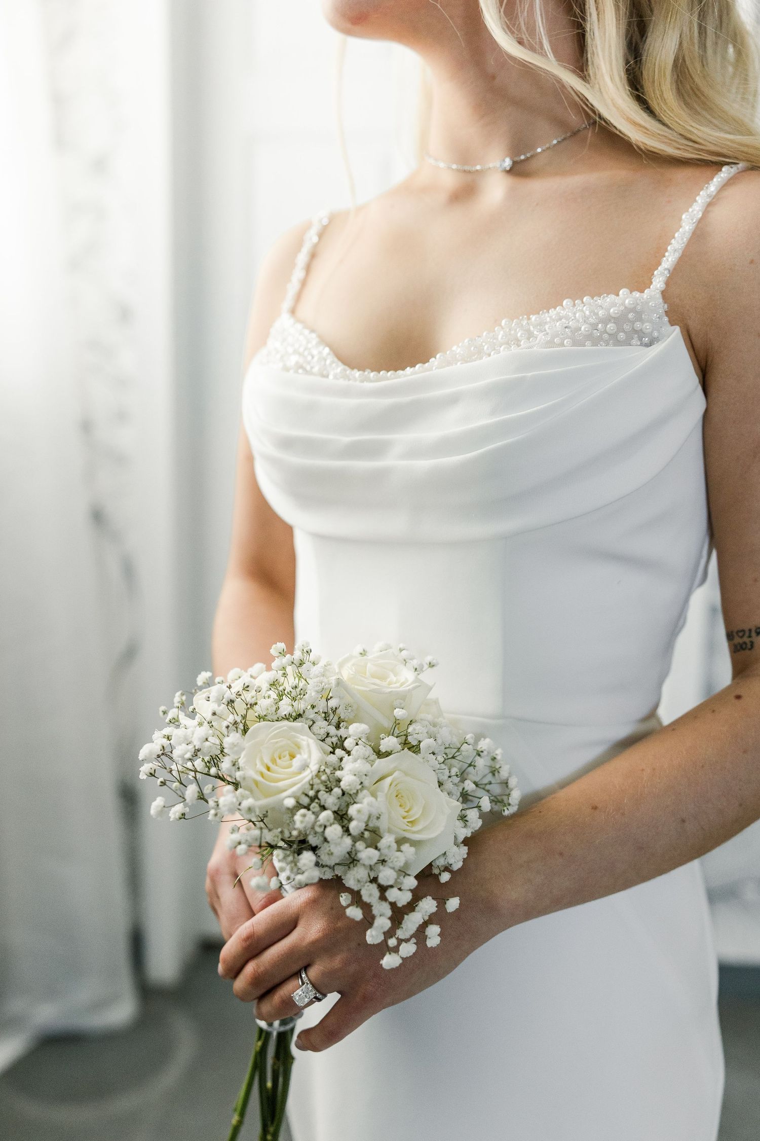 Bride in white spaghetti strap gown holding white rose and baby's breath bouquet, wearing diamond ring and pearl necklace.