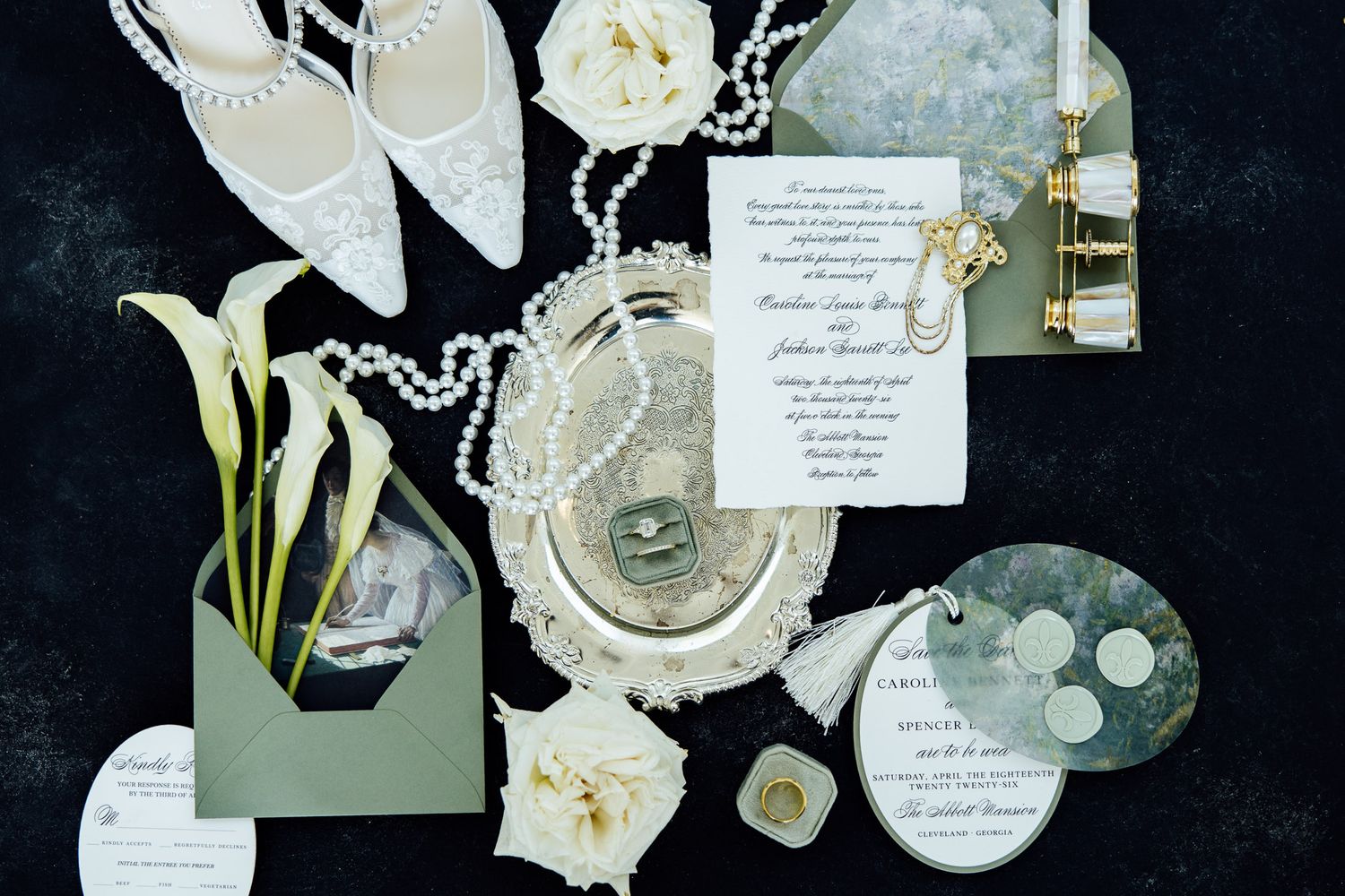 Elegant wedding flat lay with pearl necklace, lace heels, calla lilies, invitations, and vintage accessories on dark background.