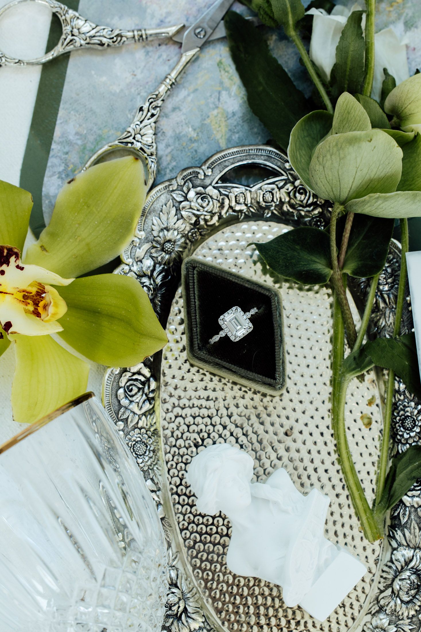 Elegant diamond engagement ring in black velvet box on ornate silver tray with green orchids and eucalyptus.
