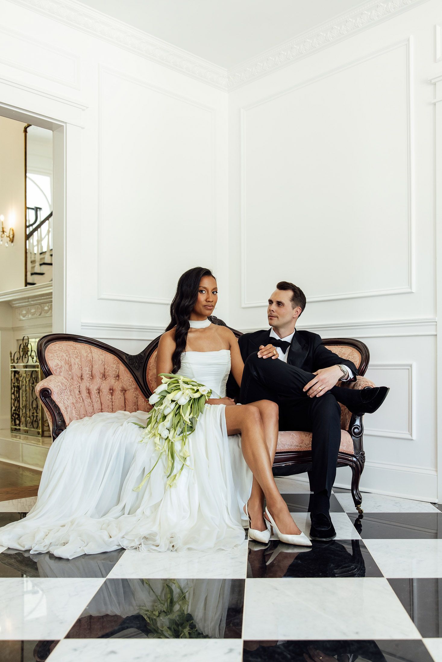 Bride and groom sitting on a vintage pink sofa in an elegant white room with black and white marble floors.