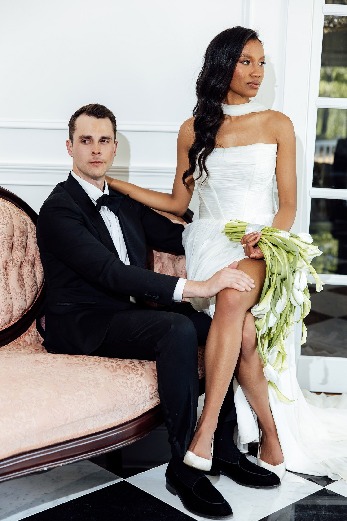 Elegant interracial wedding couple posing in luxury interior, groom in tuxedo seated on vintage sofa, bride in white dress holding bouquet.