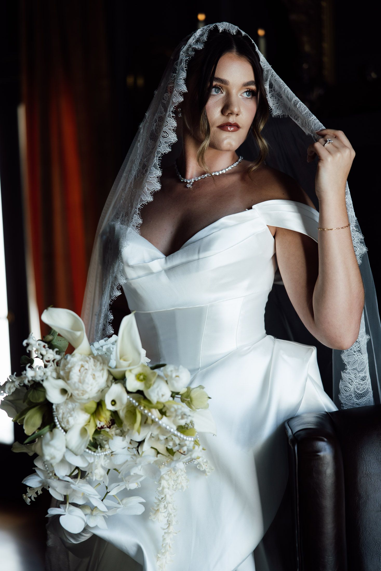 Bride in off-shoulder white gown with lace veil holds bouquet of white flowers, seated elegantly in dark setting.