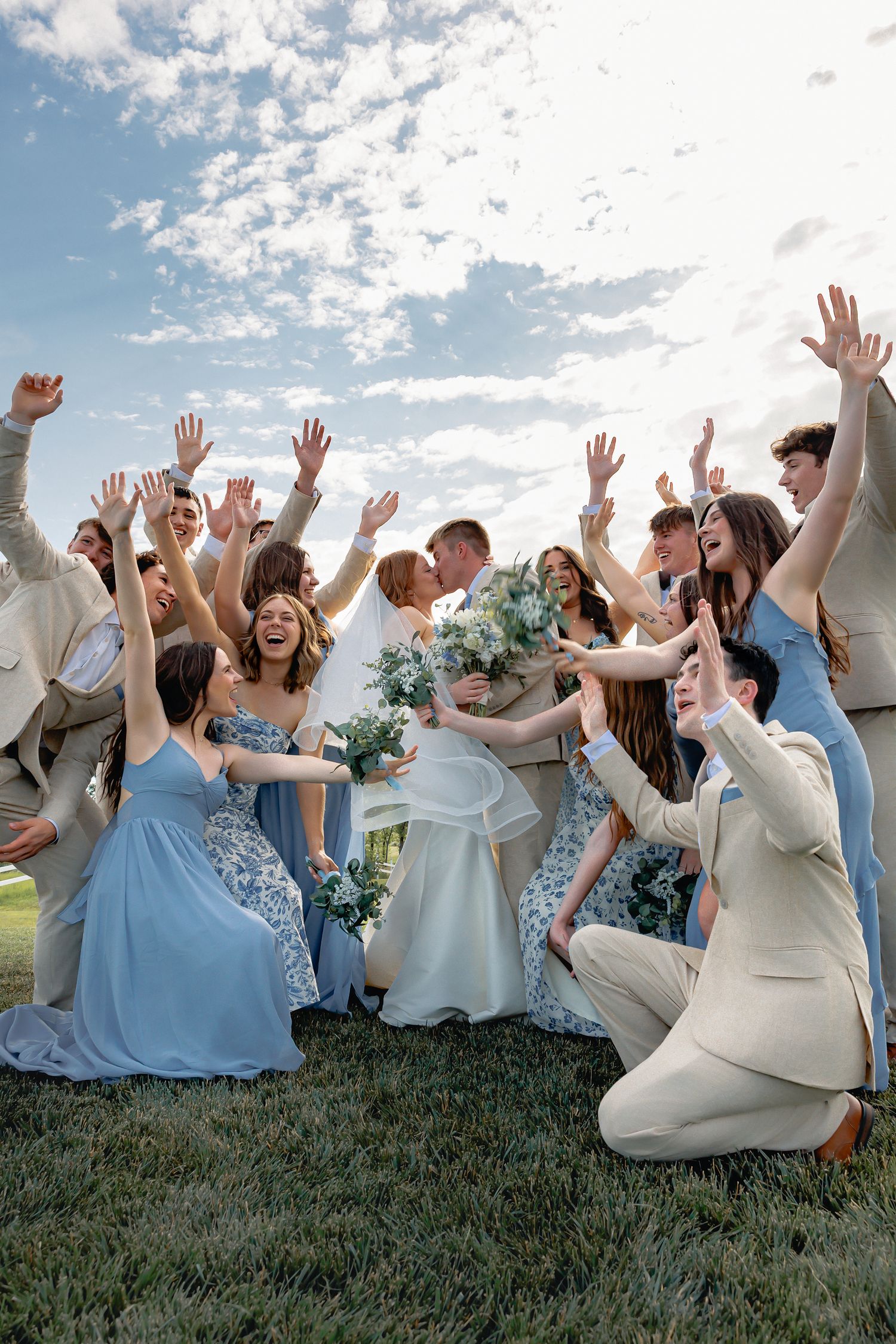 Wedding party celebrating newlyweds kissing outdoors, bridesmaids in blue dresses and groomsmen in tan suits cheering with raised arms.