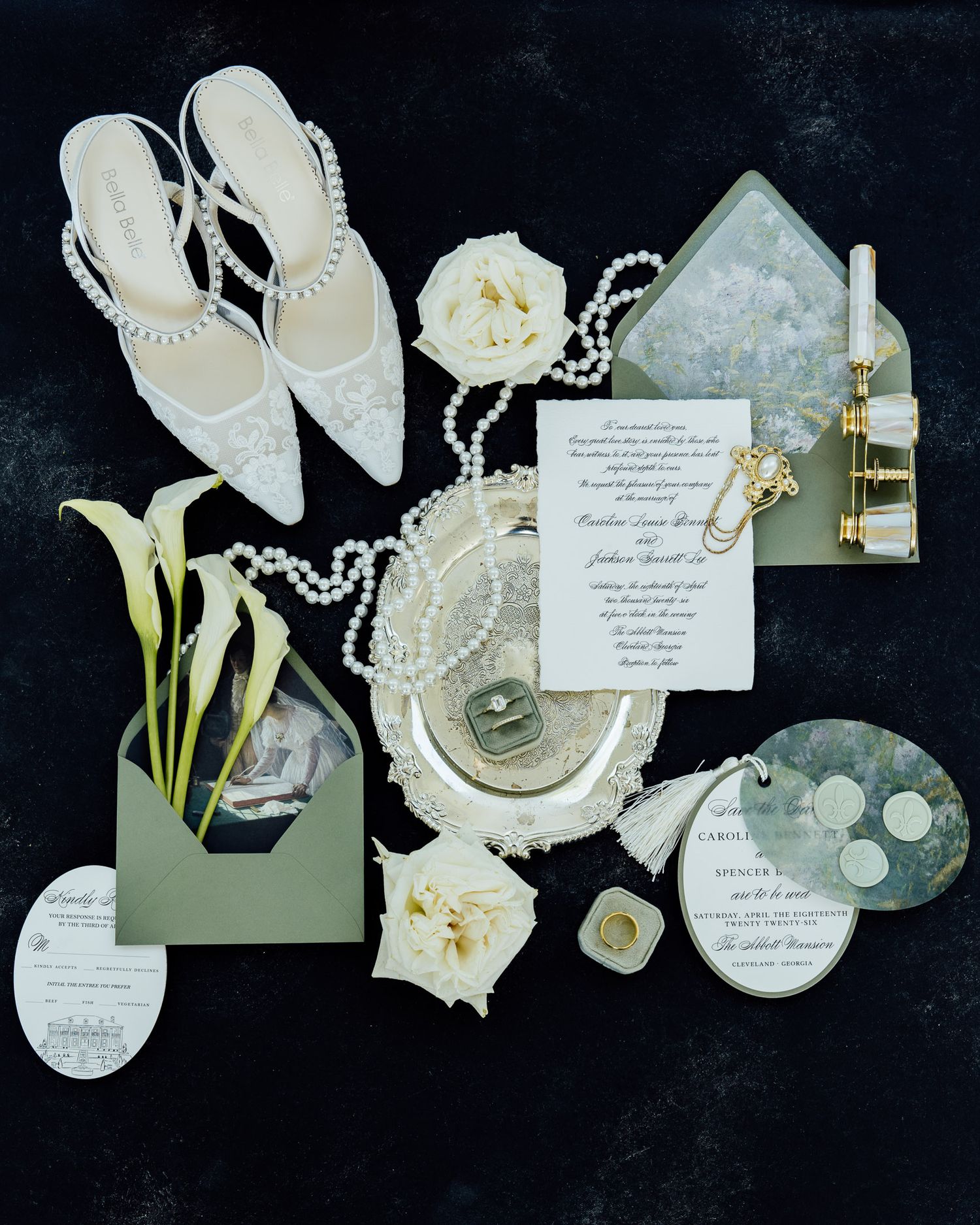 Elegant wedding flat lay with pearl heels, floral invitations, rings, calla lilies, and sage green envelopes on dark background.