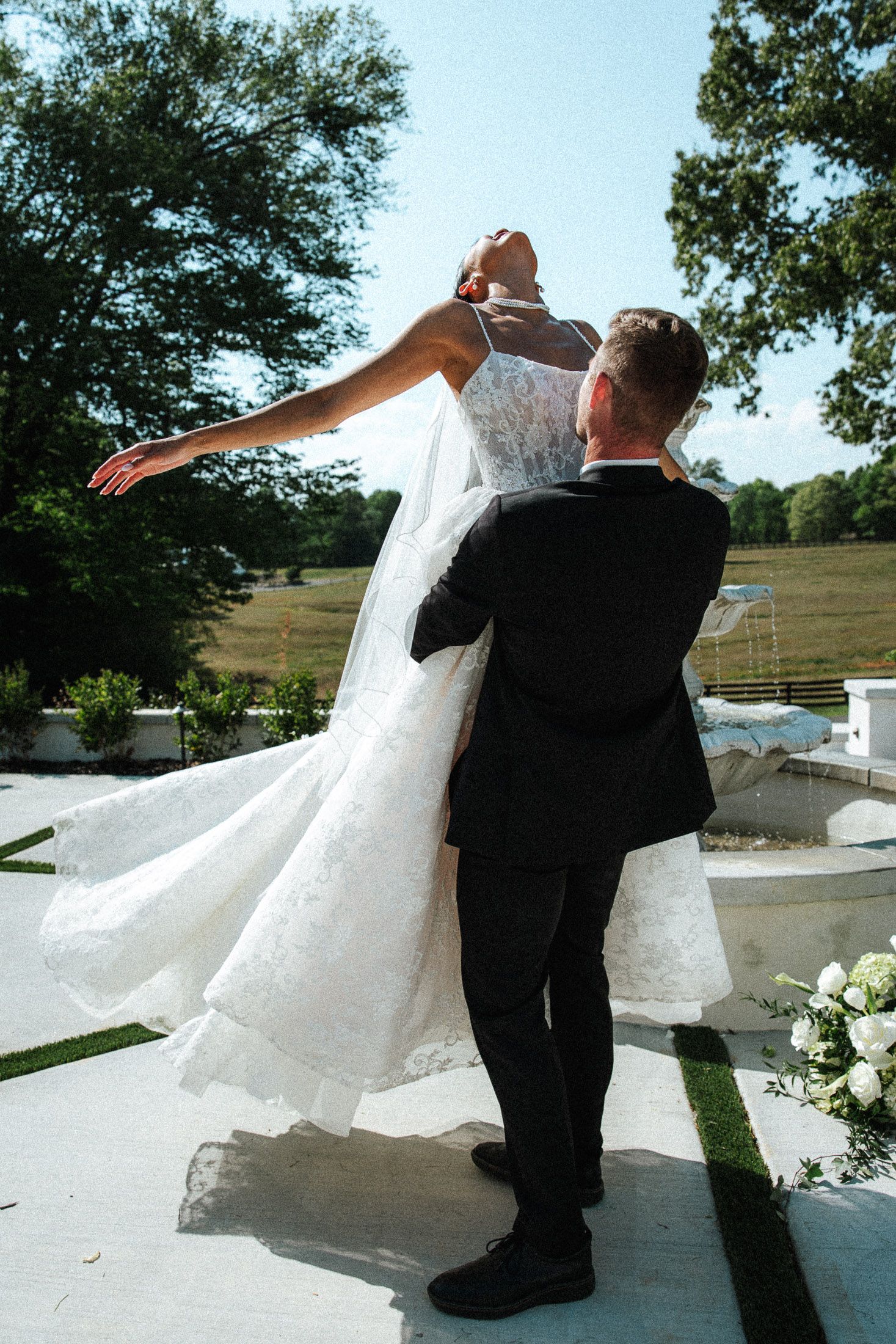 Groom lifting bride in white lace wedding dress and veil outdoors, with green trees and field in background.