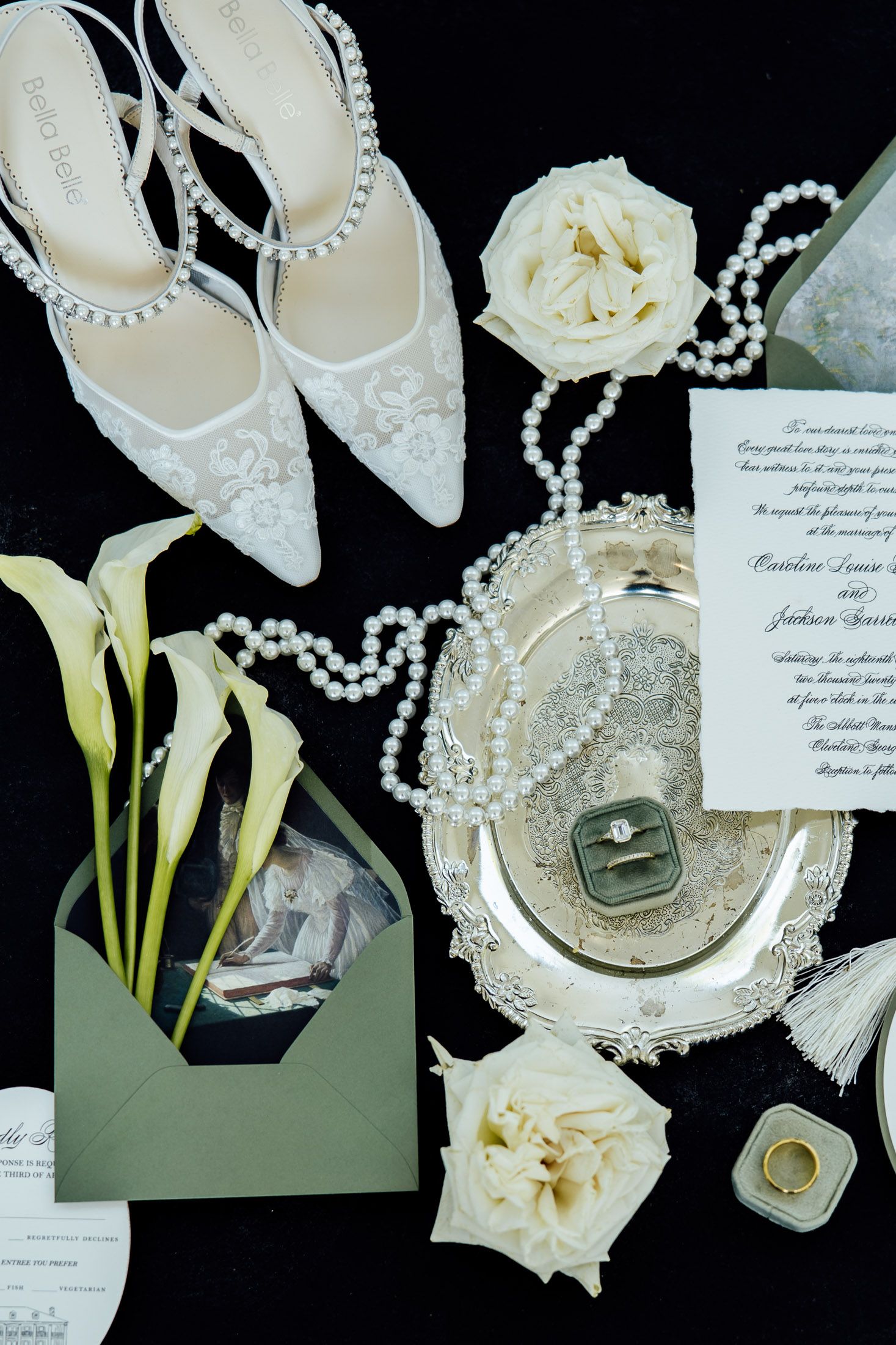Elegant bridal flat lay with white heels, pearl necklace, wedding rings, calla lilies, roses, and invitation on black background.