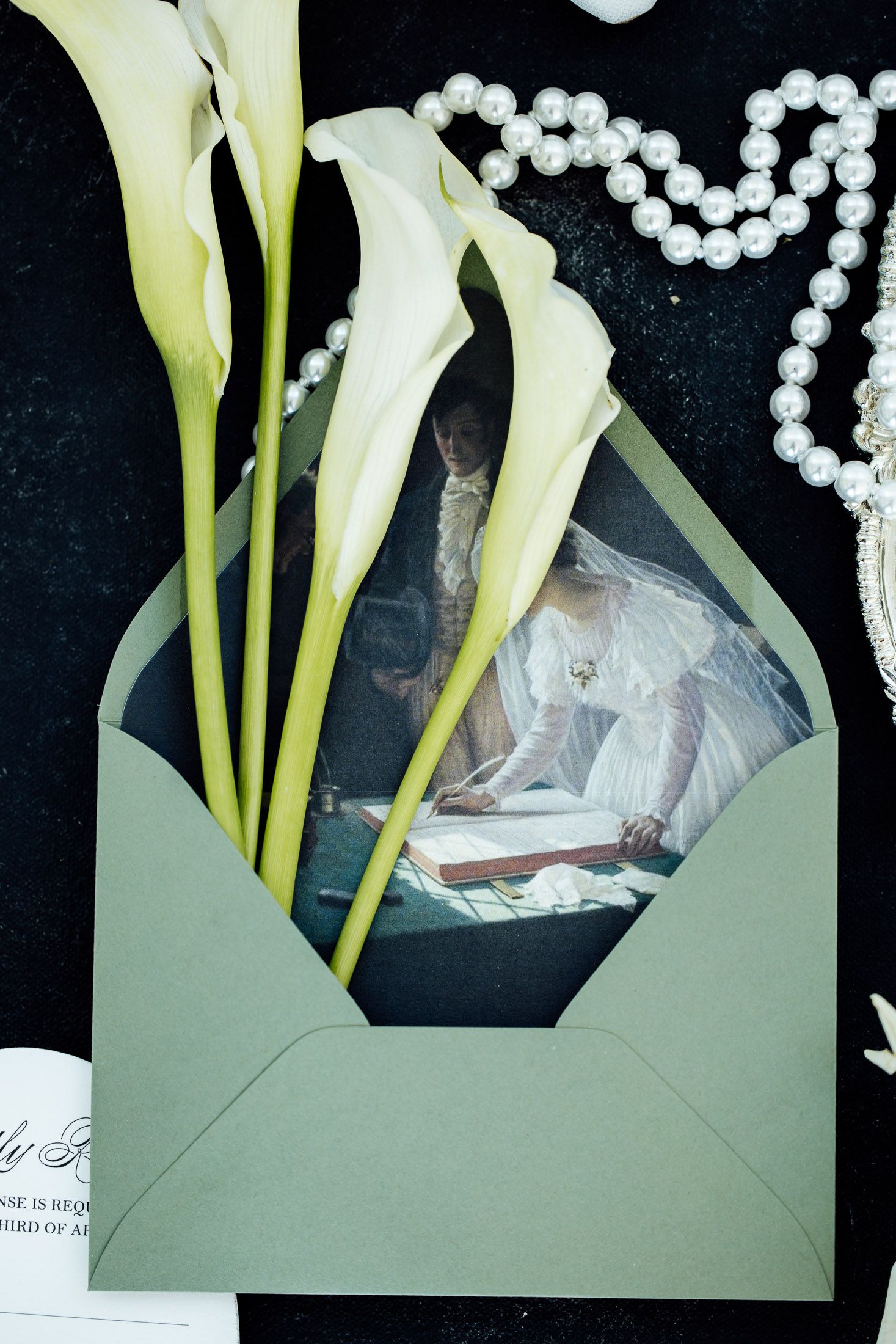 Green envelope with wedding photo, white calla lilies, and pearl necklace arranged on dark background for bridal flat lay.