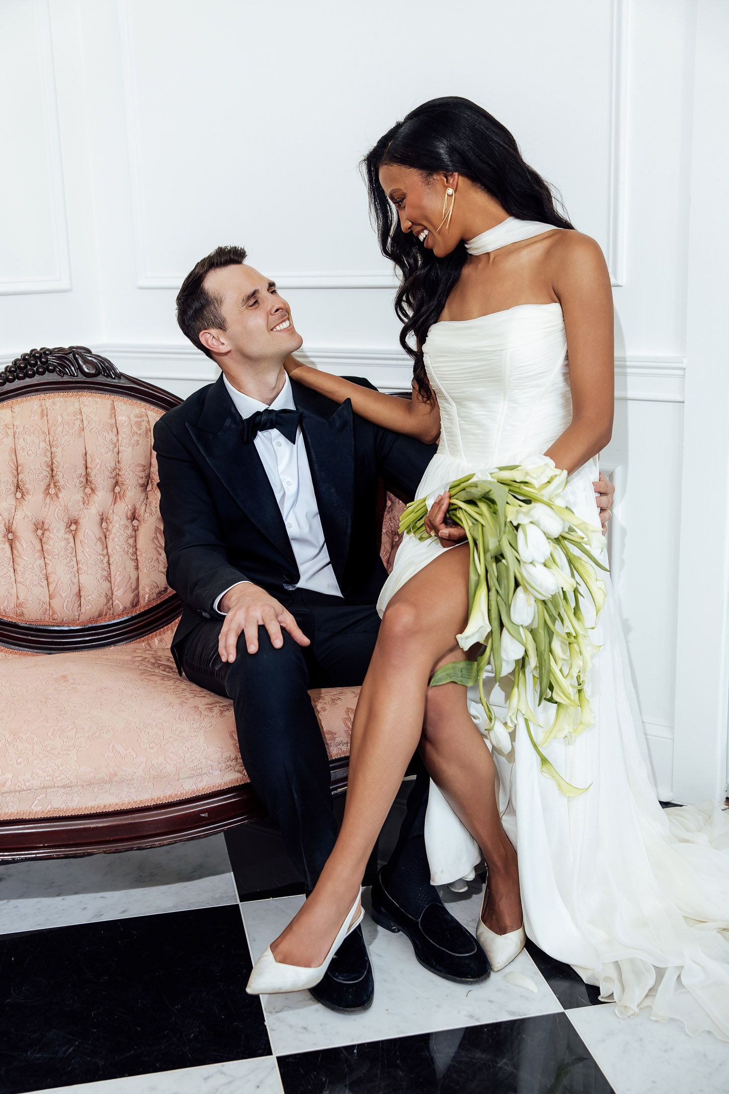 Elegant interracial wedding couple, groom in tuxedo seated on vintage sofa, bride in white dress holding tulip bouquet.