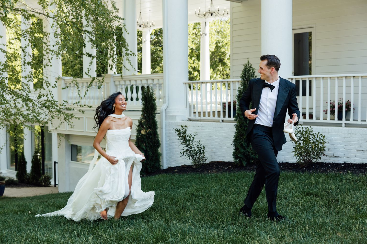 Bride and groom laughing and running on green lawn outside elegant white colonial wedding venue with columns.
