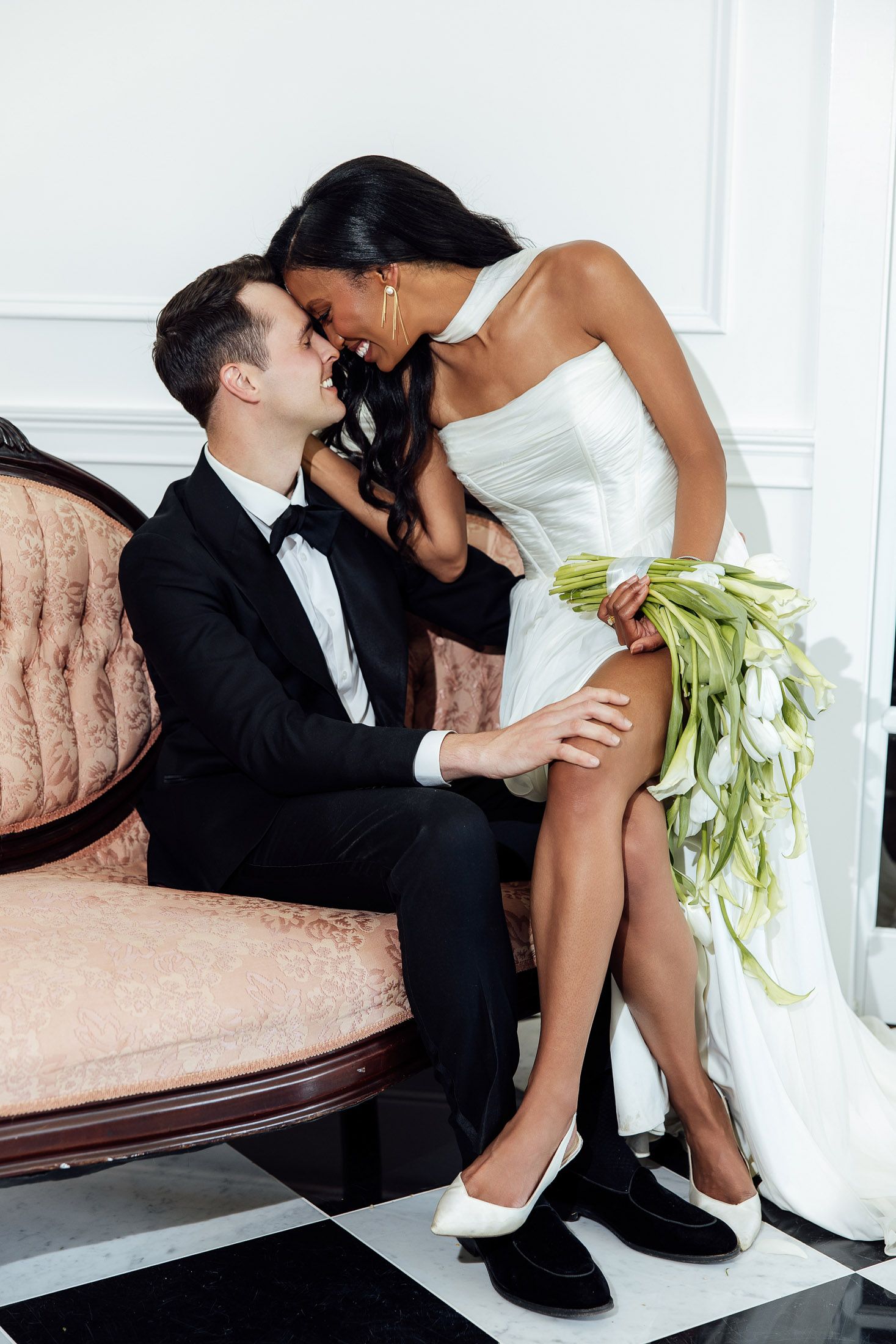 Elegant interracial couple in wedding attire sharing a romantic moment on a vintage pink sofa with white tulip bouquet.